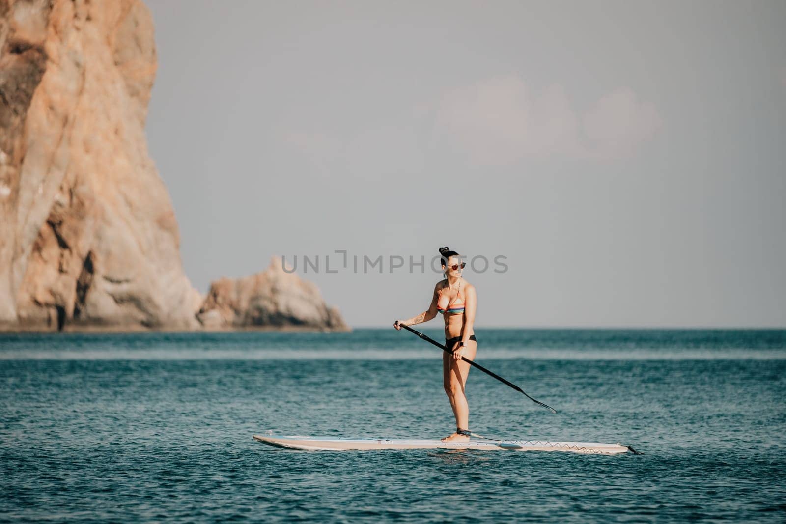 Sea woman sup. Silhouette of happy middle aged woman in rainbow bikini, surfing on SUP board, confident paddling through water surface. Idyllic sunset. Active lifestyle at sea or river