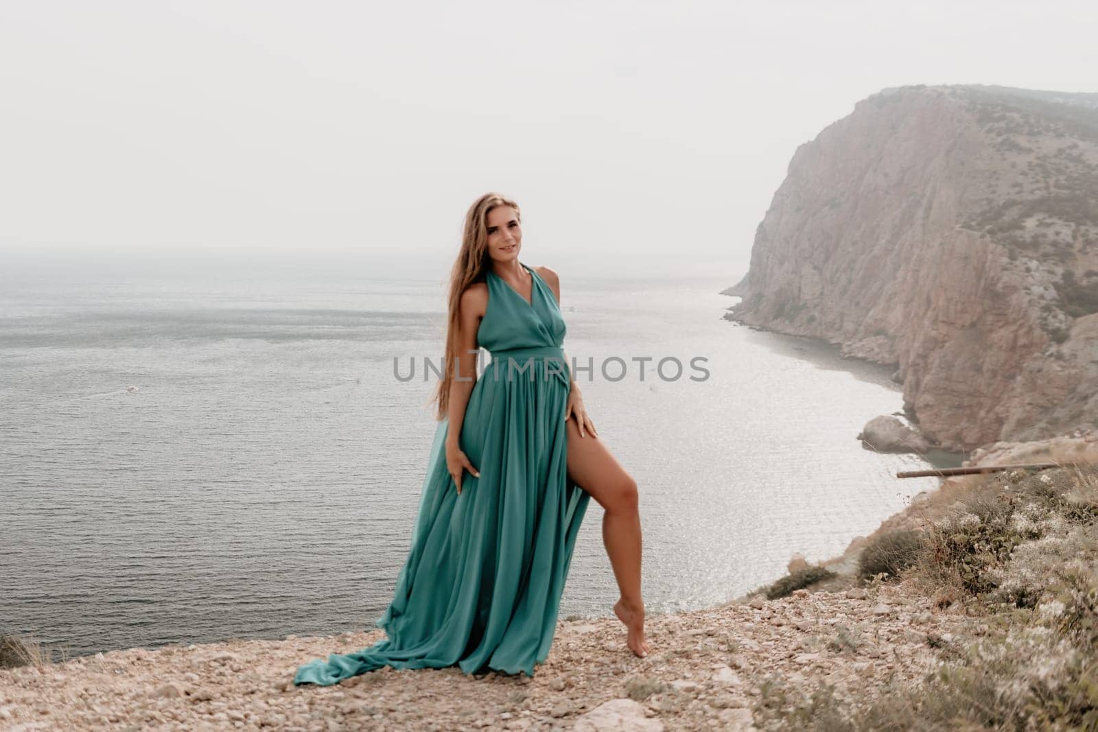 Side view a Young beautiful sensual woman in a mint long dress posing on a volcanic rock high above the sea during sunset. Girl on the nature on overcast sky background. Fashion photo