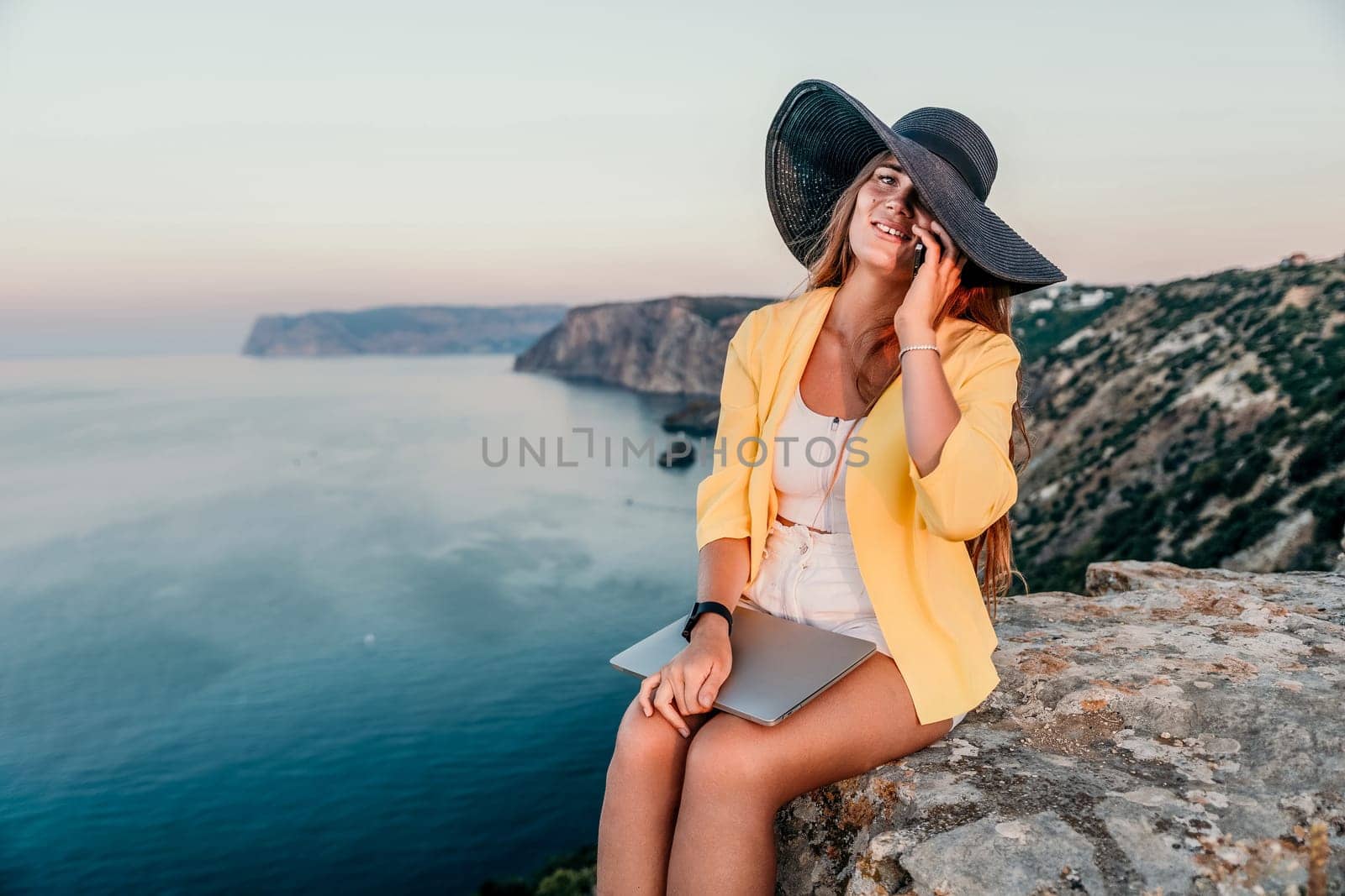 Successful business woman in yellow hat working on laptop by the sea. Pretty lady typing on computer at summer day outdoors. Freelance, travel and holidays concept.