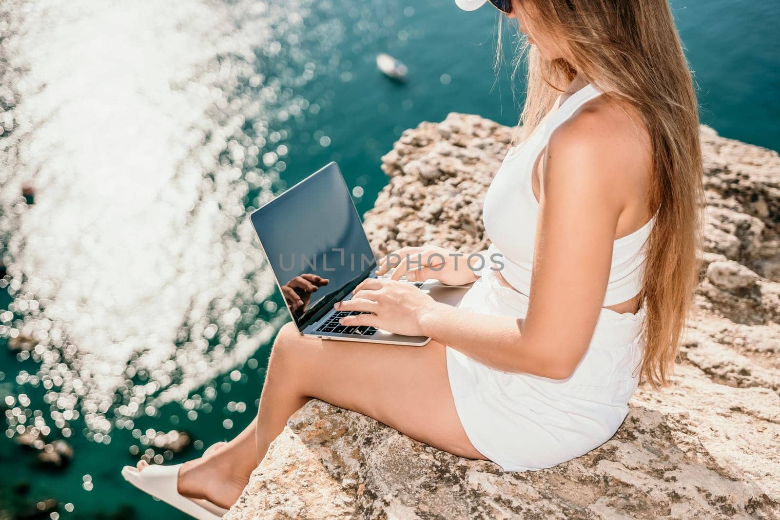 Successful business woman in yellow hat working on laptop by the sea. Pretty lady typing on computer at summer day outdoors. Freelance, travel and holidays concept.