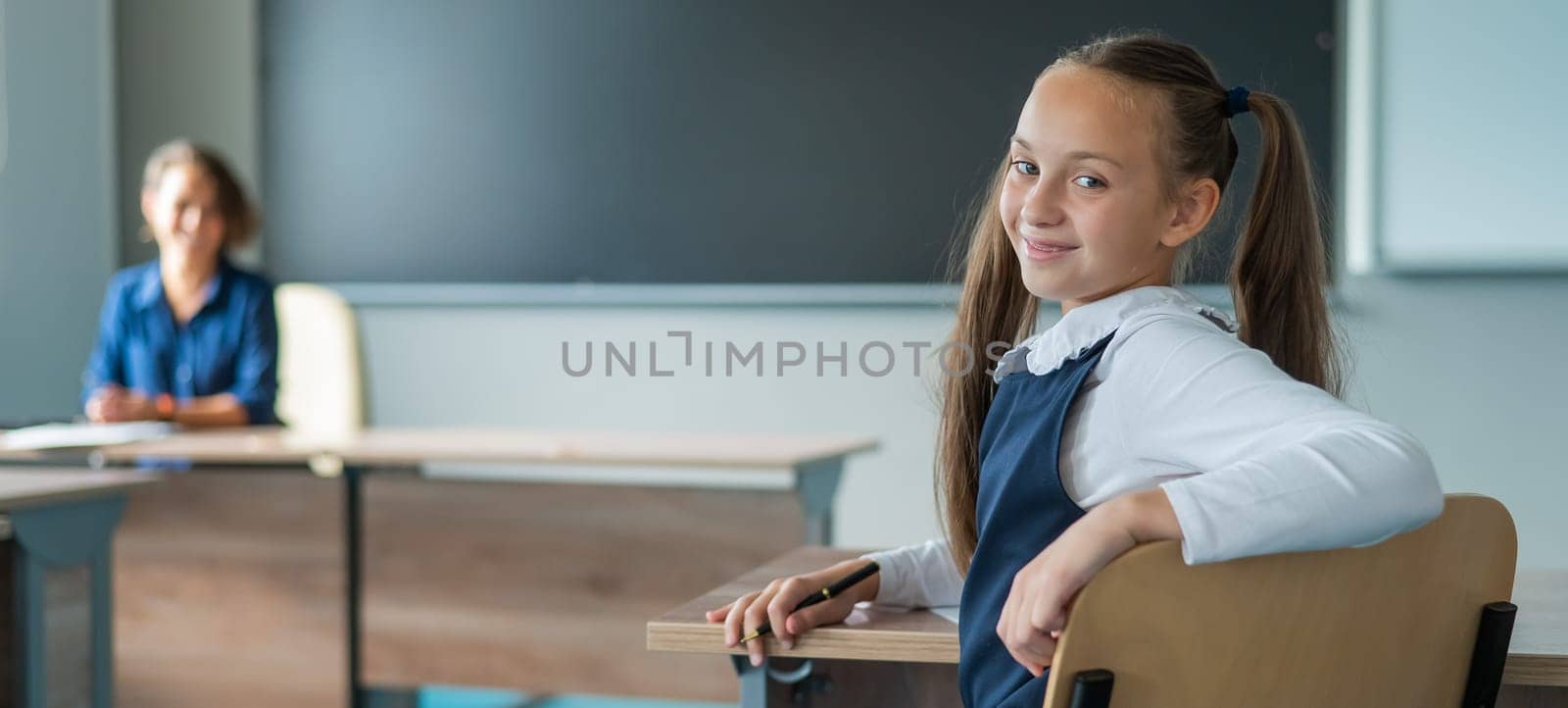 Caucasian girl and her teacher in the classroom. Schoolgirl turns around and looks at the camera