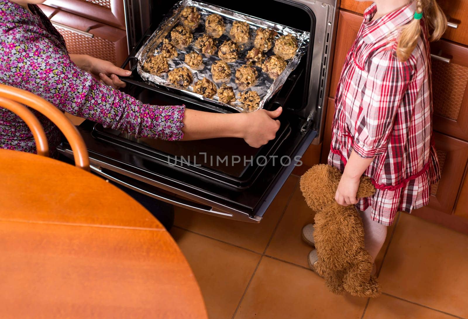 Happy mother and daughter taking freshly baked Christmas cookies from oven while cooking together in cozy kitchen during winter holidays, little girl helping mom to prepare for New Year