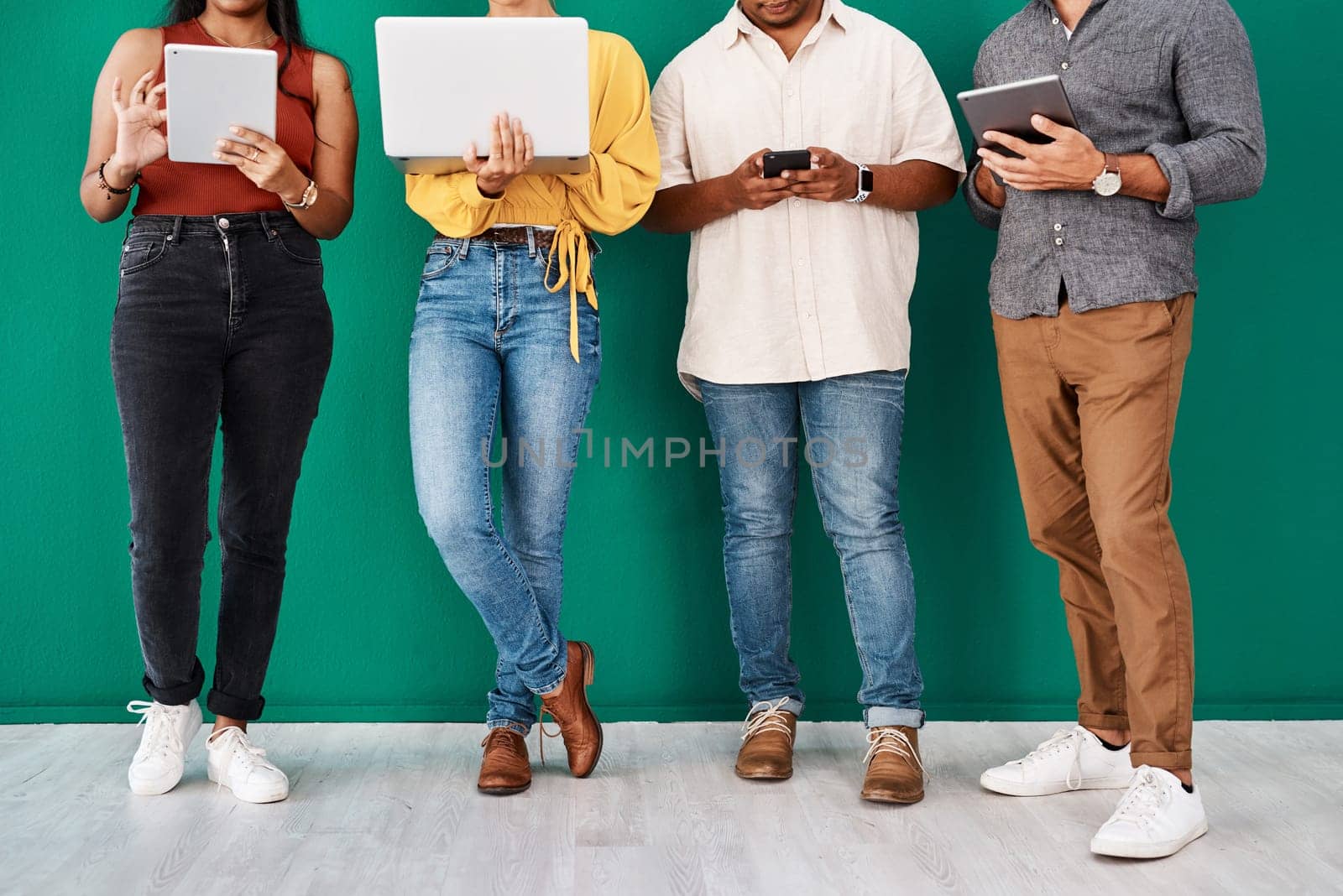There are many ways to stay connected. Closeup shot of a group of unrecognisable designers using digital devices while standing together against a green background. by YuriArcurs