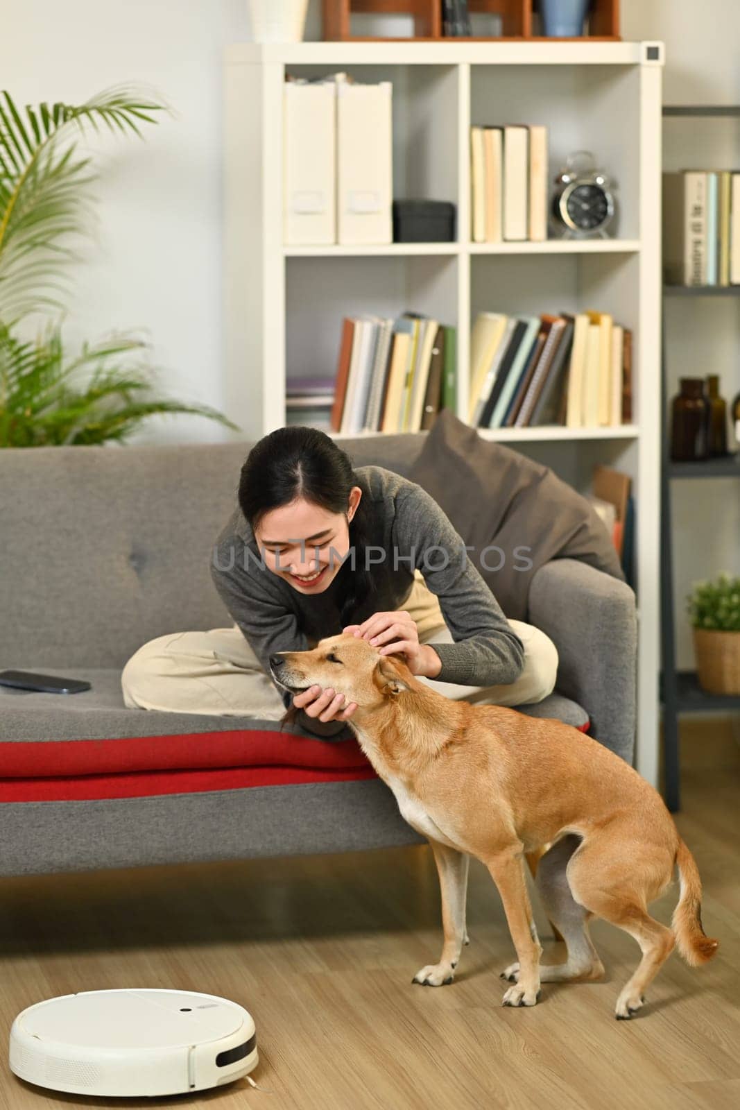 Young woman and dog sitting in living room with robot vacuum cleaner cleaning the floor. Smart home and automatic cleaning concept by prathanchorruangsak