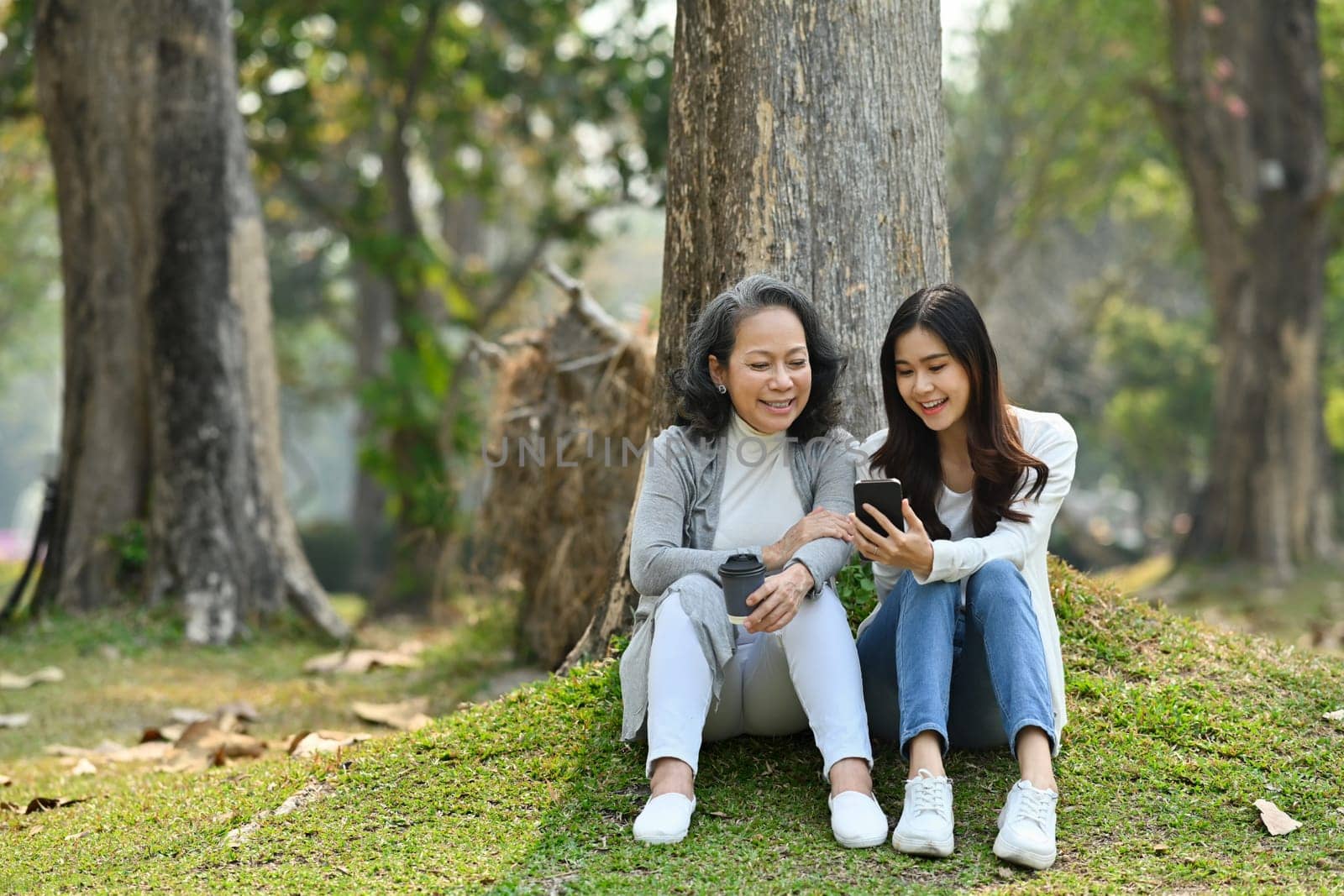 Happy elderly woman and daughter using mobile phone, enjoy outdoor leisure activity in calm nature park.