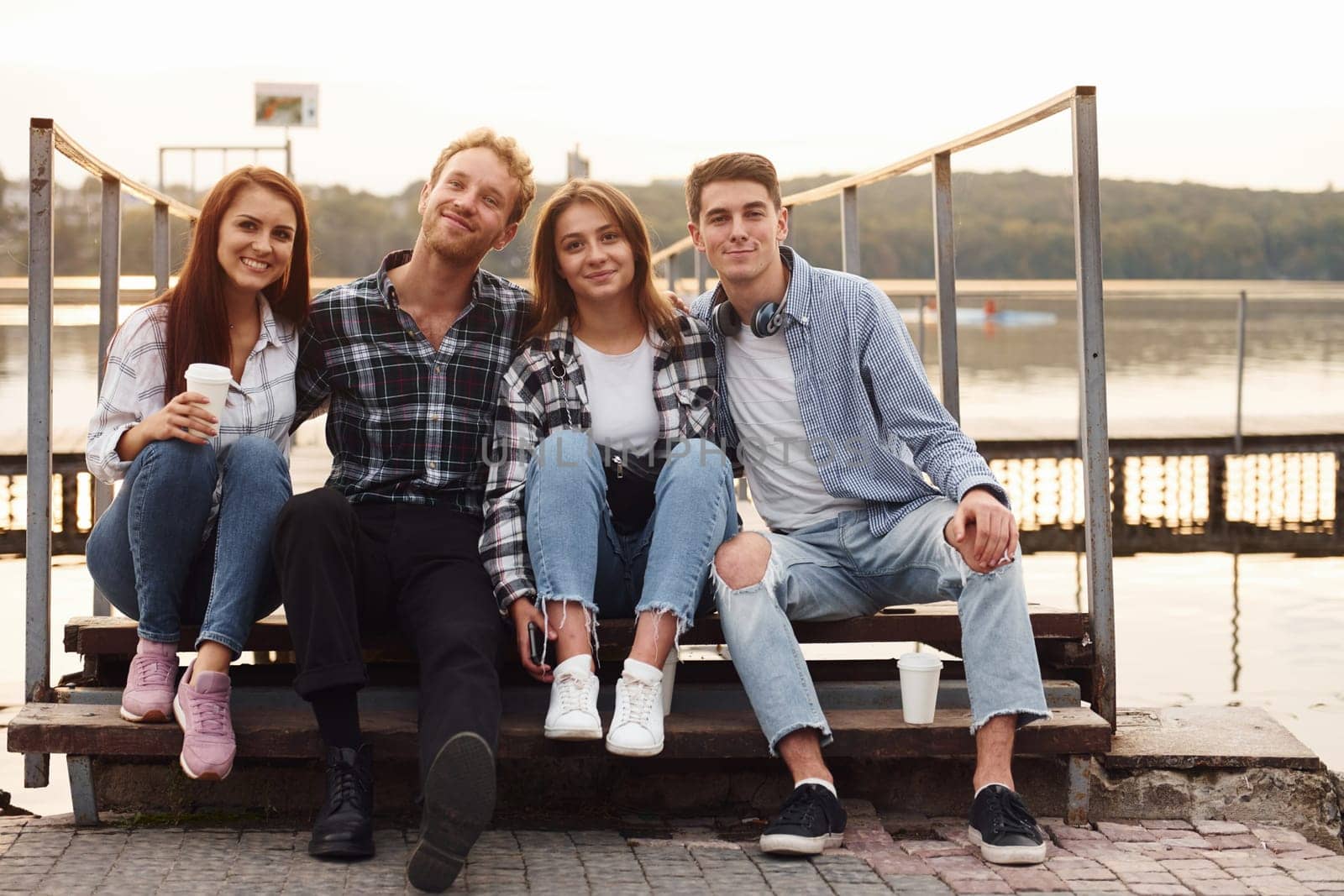 Sitting near lake and embracing each other. Group of young cheerful friends that is outdoors having fun together.