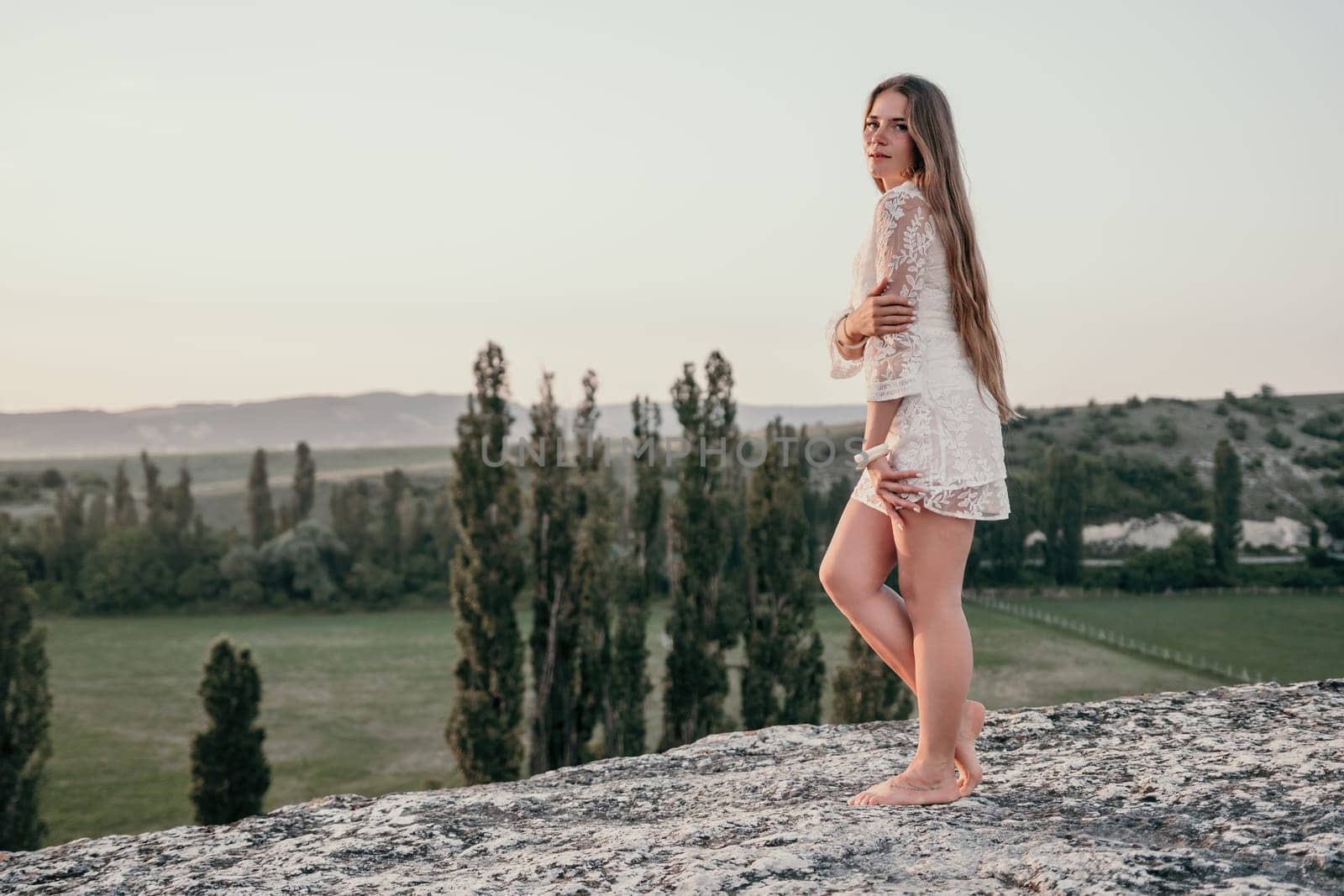 Romantic beautiful bride in white dress posing with sea and mountains in background. Stylish bride standing back on beautiful landscape of sea and mountains on sunset