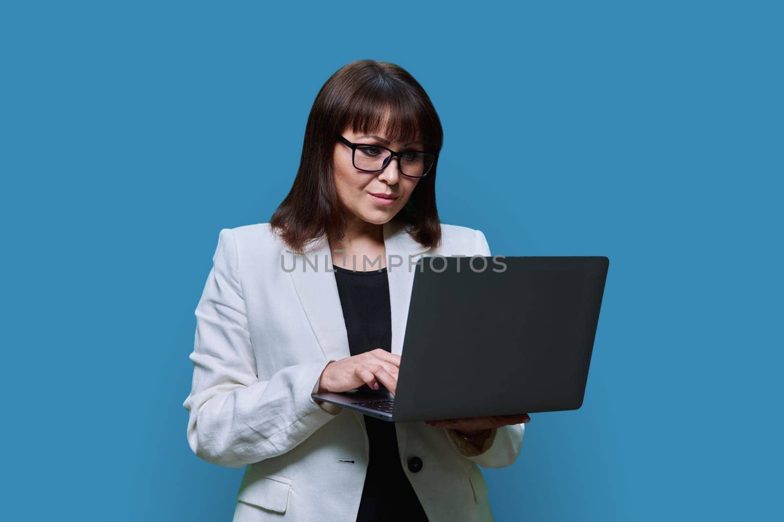 Mature business woman using laptop, on blue studio background. Confident serious middle aged female in glasses suit looking at screen. Technology businessperson management teaching finance insurance