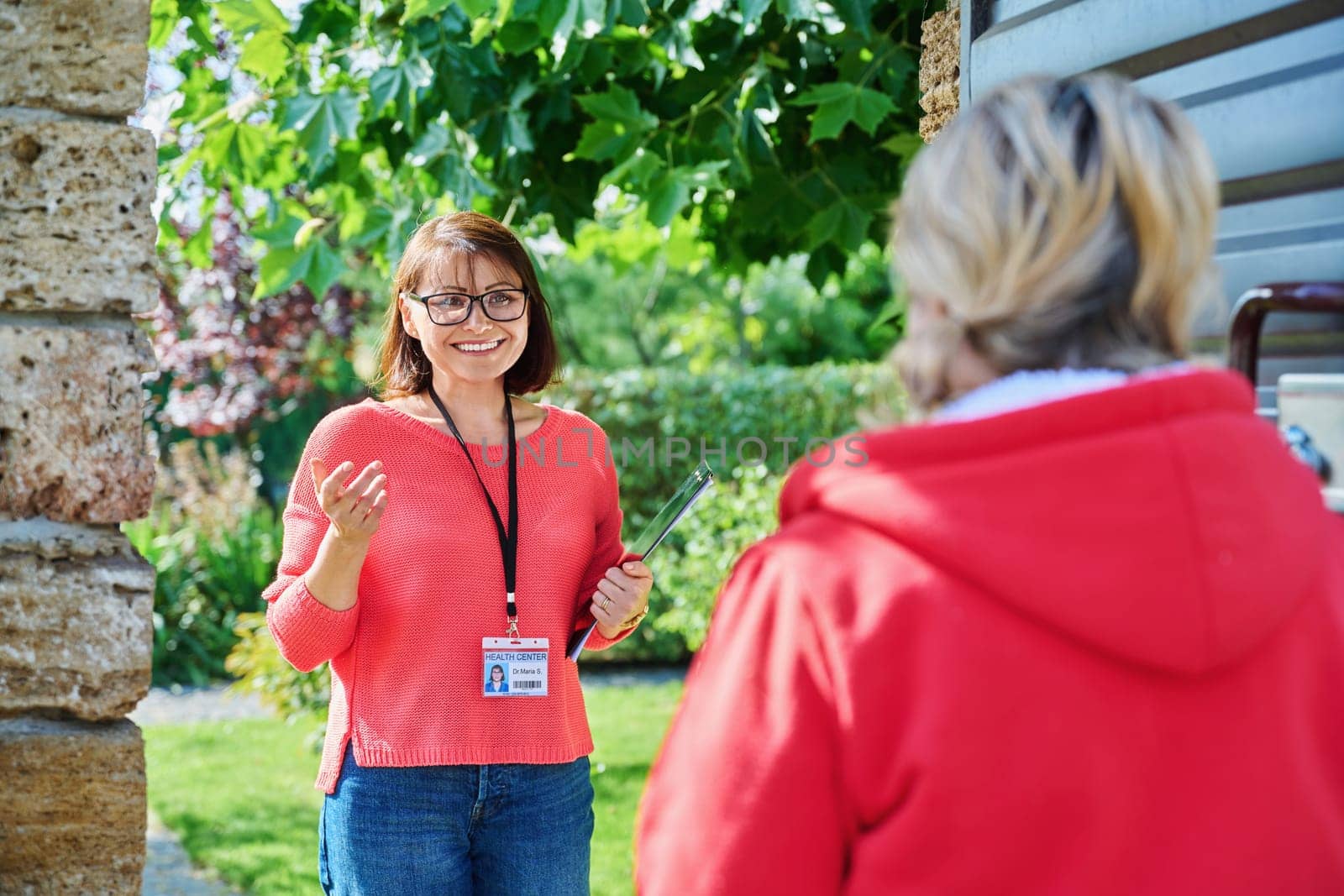 Female social counselor, health center worker, meets middle-aged woman near entrance gate to private backyard area