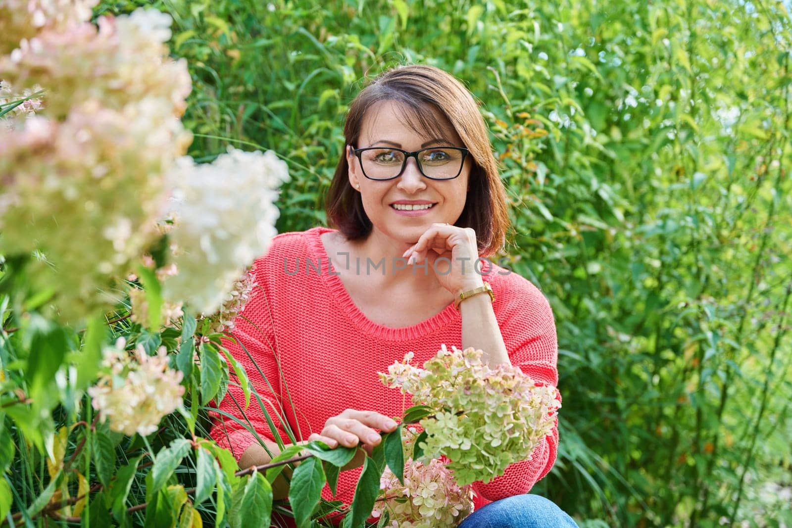 Portrait of smiling middle aged woman looking at camera outdoor. Successful confident mature woman in red, nature green lawn background