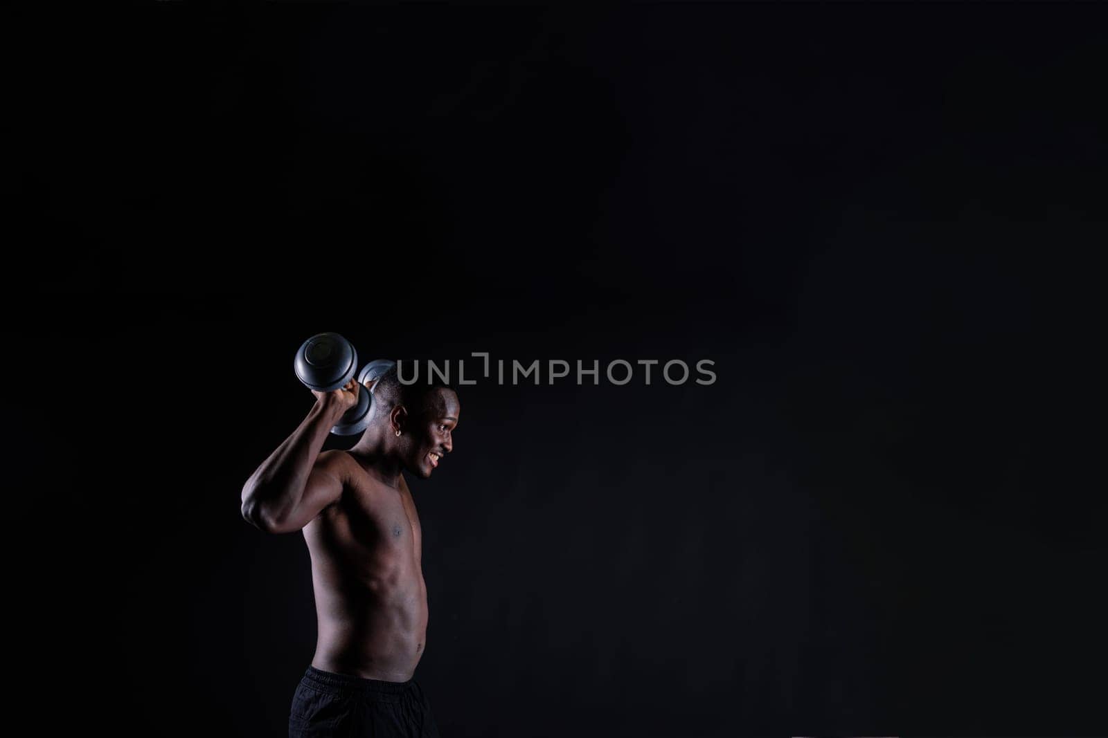 Confident young man shirtless portrait training with dumb-bells against black background. by Zelenin