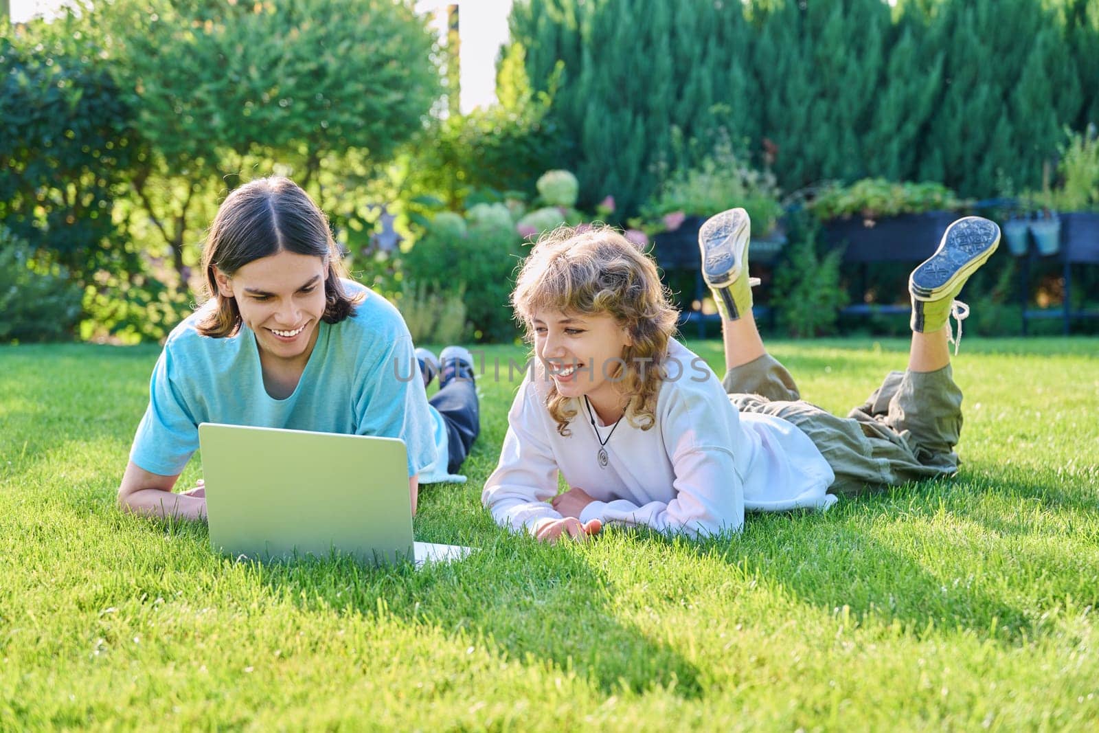 Two teenage friends of students lying on grass with laptop, in backyard, guy and girl 16, 17 years old study together. Friendship, youth, technology, high school, college, lifestyle concept