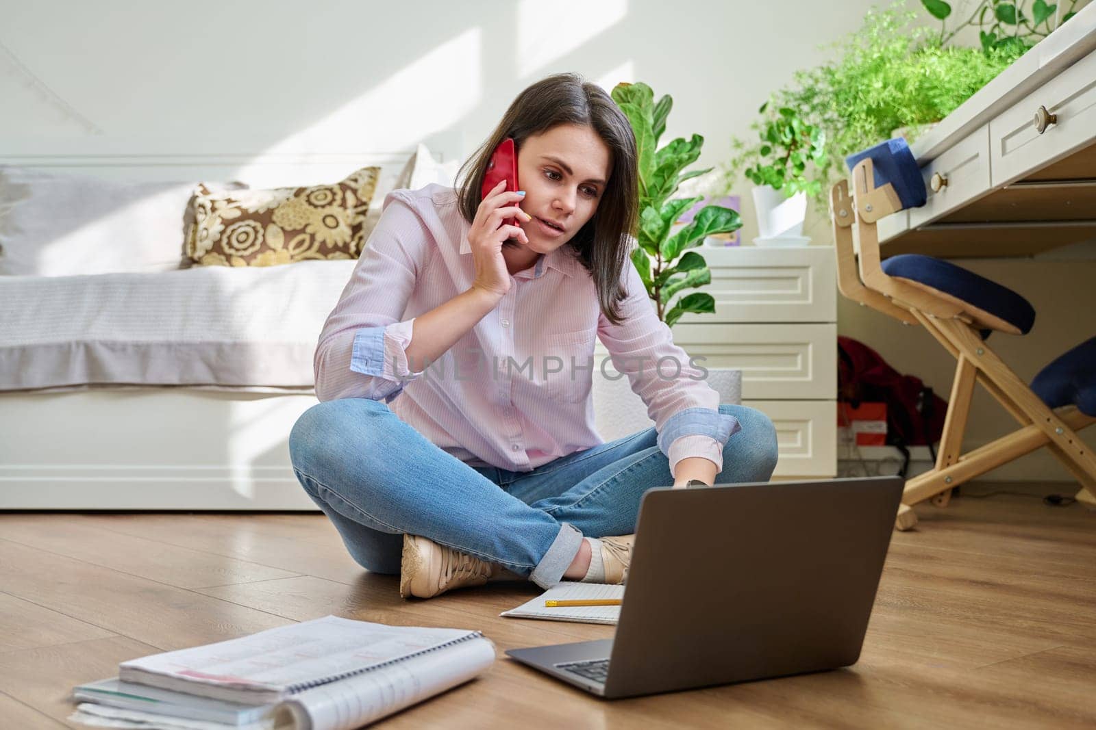 Young female university student studying at home sitting on floor using laptop phone. E-learning, e-education, self-learning, remote lessons, technology, knowledge, education concept