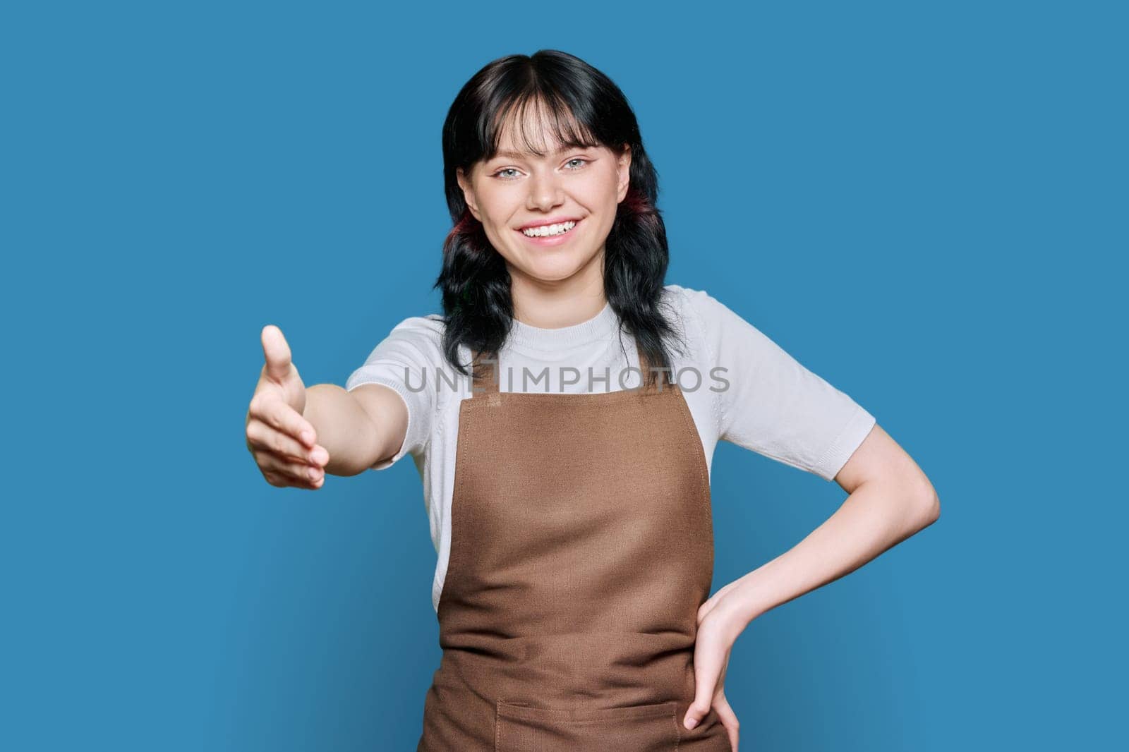 Young woman worker in an apron with a hand for a handshake, on a blue color studio background. Cooperation, partnership, business, service, staff concept