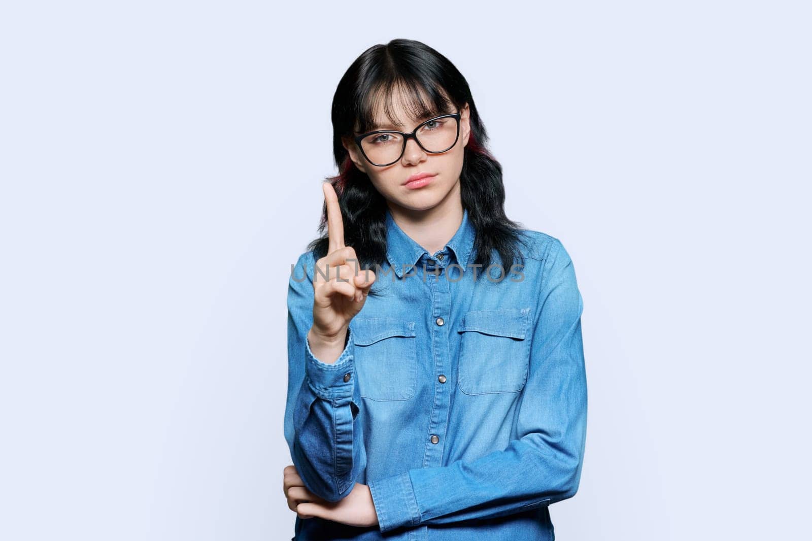 Young female student showing index finger up, over white studio background. Gesture attention idea