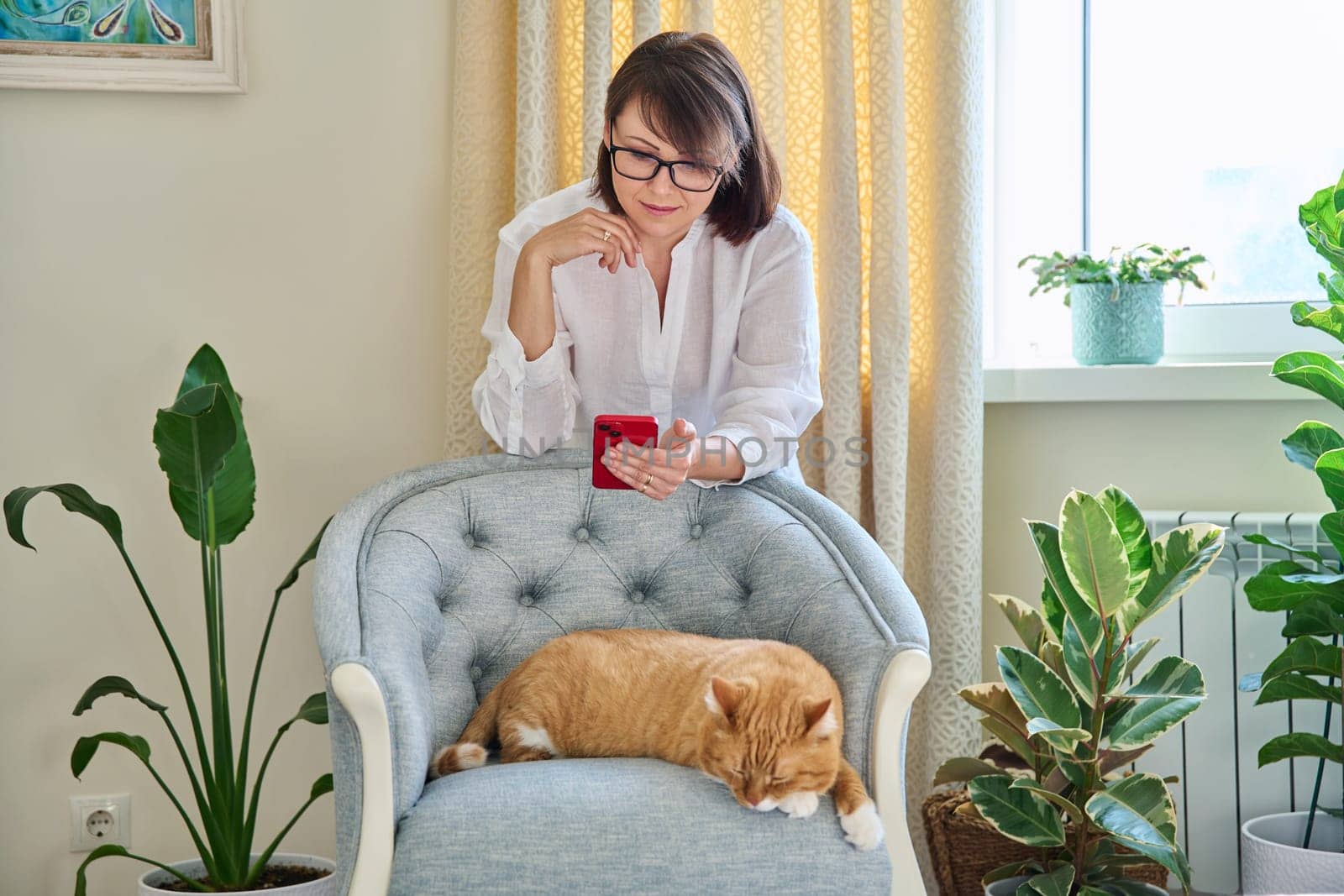 Middle aged woman using smartphone, in home interior with sleeping cat on armchair, houseplants. Lifestyle, comfort, green pets people animals concept