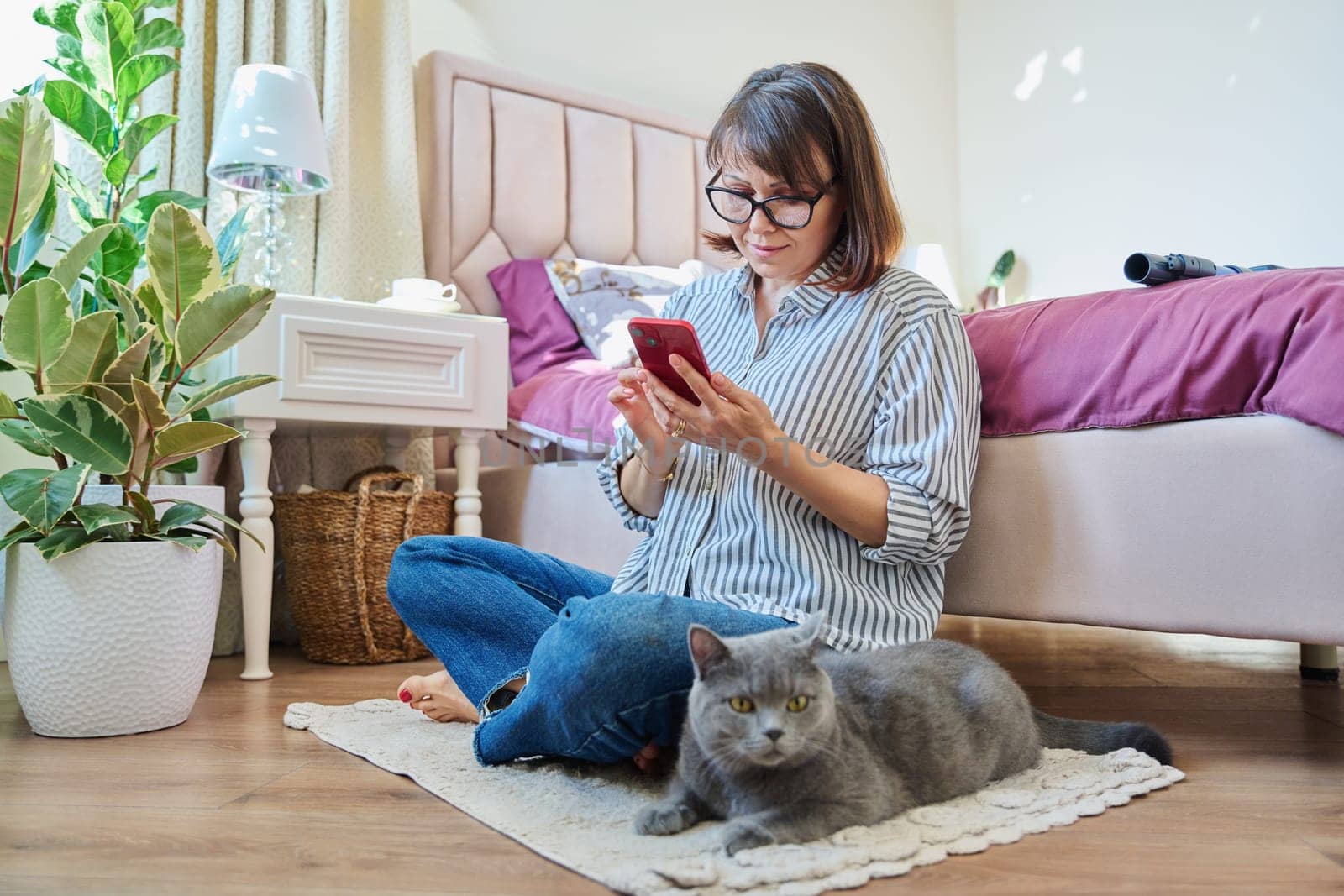 Home lifestyle, woman with cat, comfort calmness concept. Female sitting on floor on carpet using smartphone, pet gray british cat lying near owner