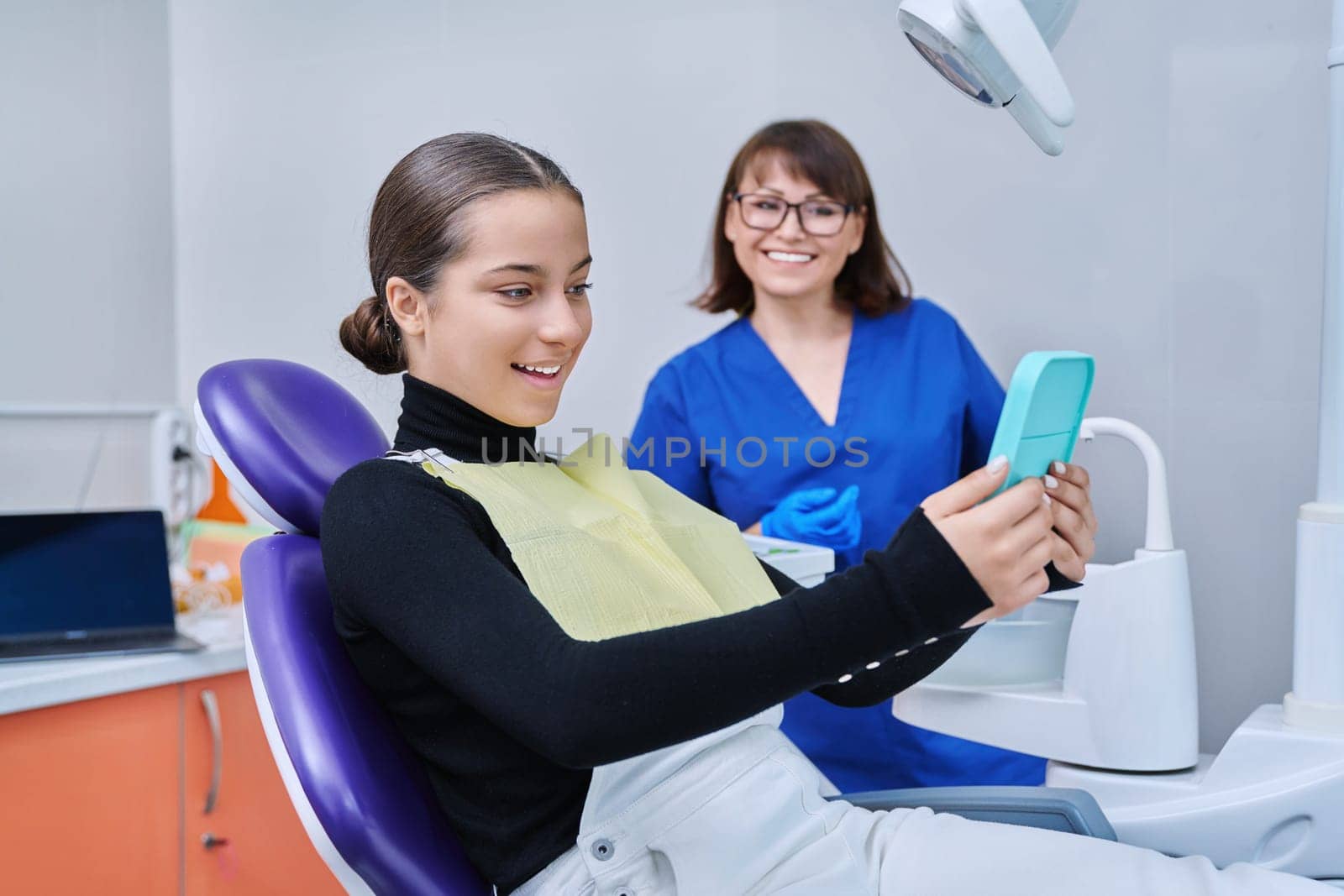 Happy female with mirror sitting in dentist chair looking at healthy teeth by VH-studio