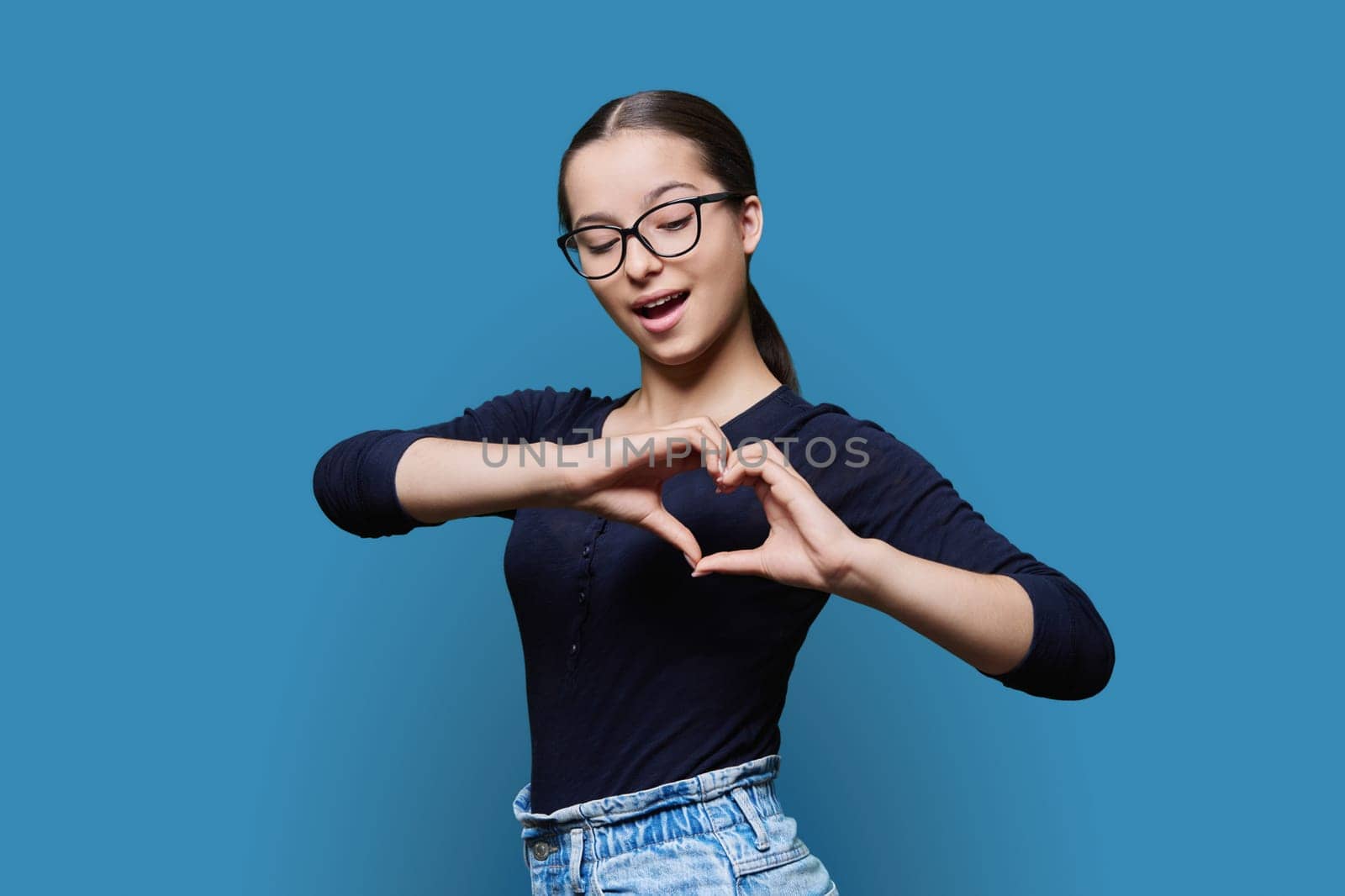 Young smiling female showing heart gesture with fingers, happy female showing love, blue studio background. Body language, signs symbols, love, emotions, romance youth concept