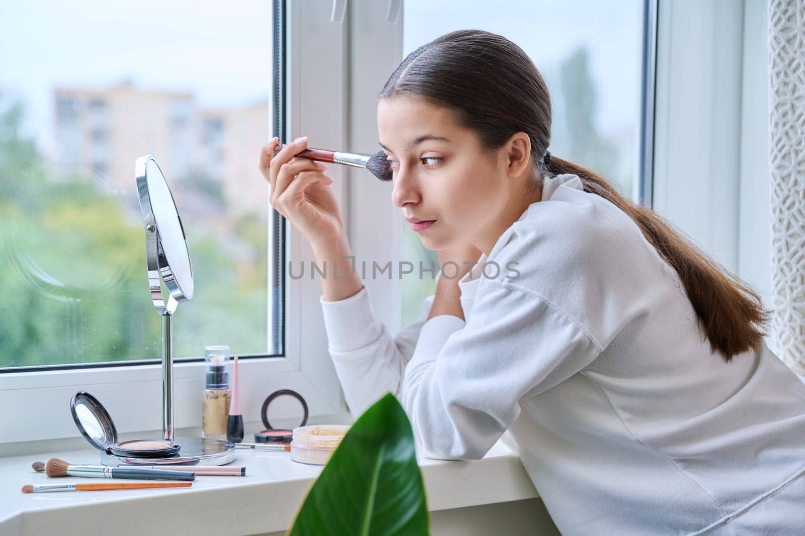 Young teenage girl with cosmetics mirror doing natural makeup, at home near the window, using brushes, cosmetics. Beauty, fashion, youth concept