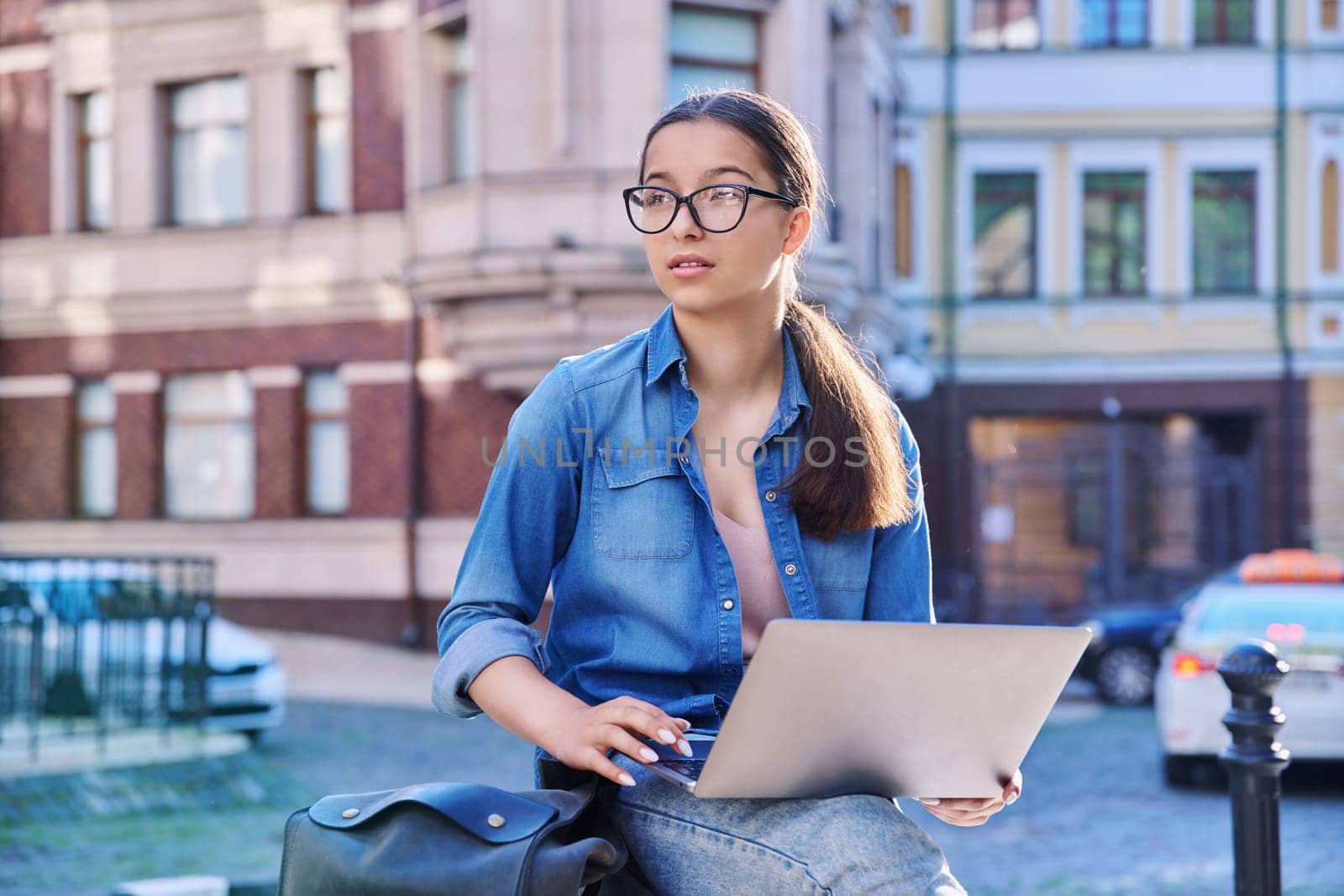 Teenage student girl in headphones using laptop outdoor, urban background. Technology, education, adolescence, lifestyle, youth concept