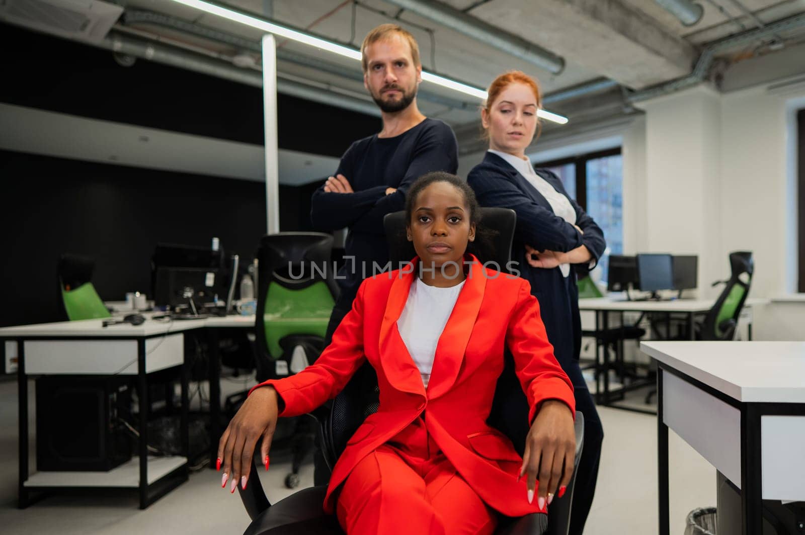 Caucasian red-haired woman, bearded Caucasian man stand behind a seated African American young woman in the office. by mrwed54