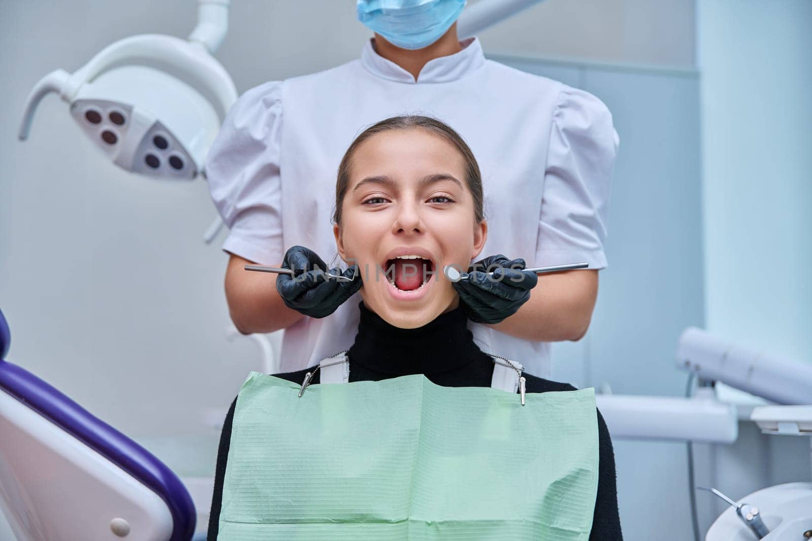 Portrait of young teenage girl in dental chair with hands of doctor with tools. Female teenager smiling with teeth looking at camera in dentist office. Adolescence hygiene treatment dental health care