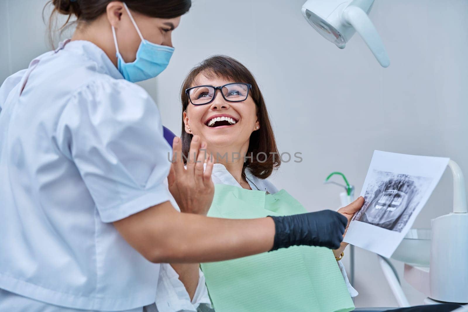 Female doctor dentist talking to middle aged woman patient in dental chair, discussing x-rays of teeth and jaws. Dental treatment and prosthetics, implantation, health care concept