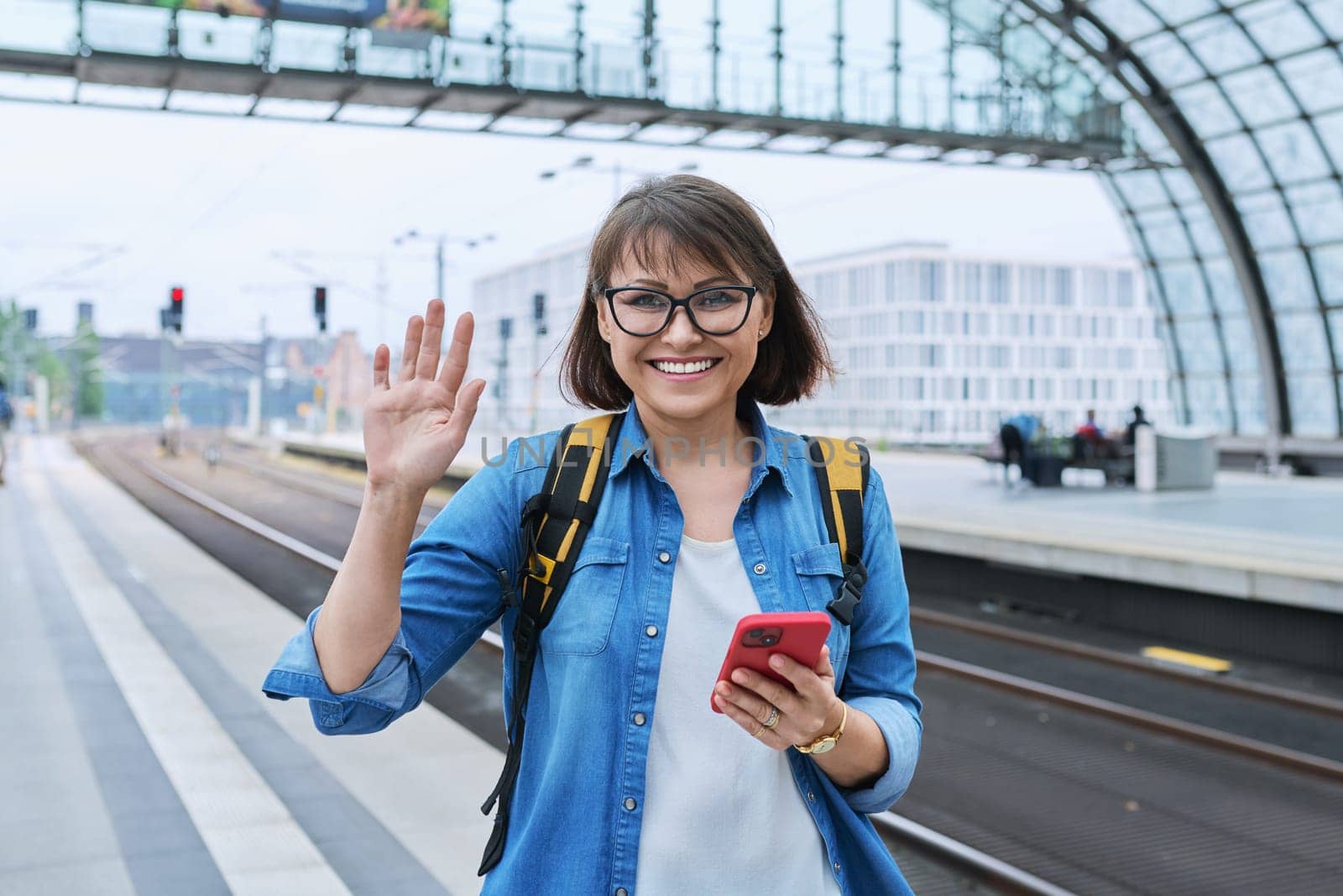 Mature happy woman with smartphone backpack looking at camera inside modern train station building, on platform. Emotion joy happiness, transportation, journey, trip, middle age people concept