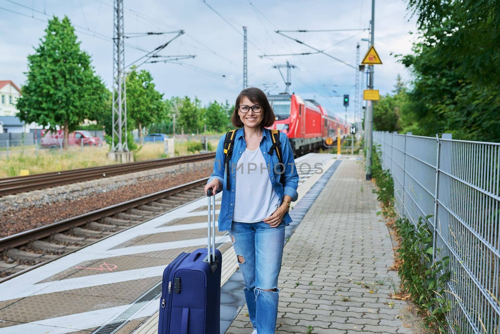 Middle aged smiling woman with suitcase backpack on outdoor platform of railway station, looking at camera, against backdrop of red electro train. Transport, journey, vacation, tourism, people concept