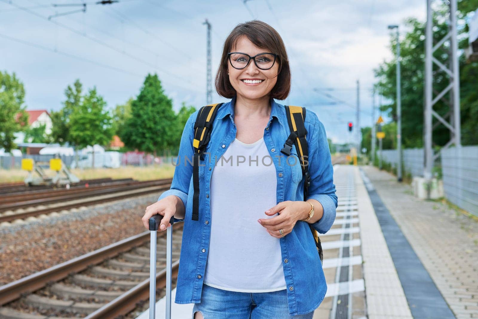 Middle aged woman with backpack with suitcase on outdoor platform of railway station looking at camera. Transport, luggage, journey, vacation, tourism, people concept
