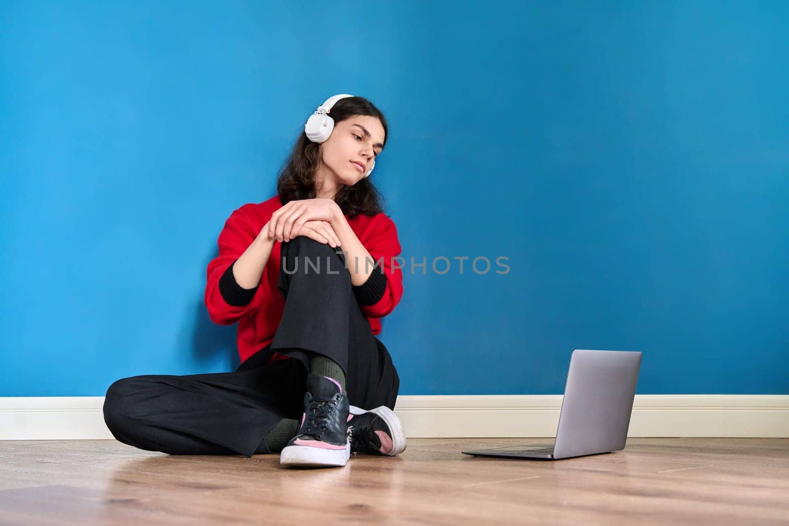 Young teenage guy student in headphones with laptop sitting on the floor on blue background. Lifestyle, youth, technology, leisure, music, education, young people concept