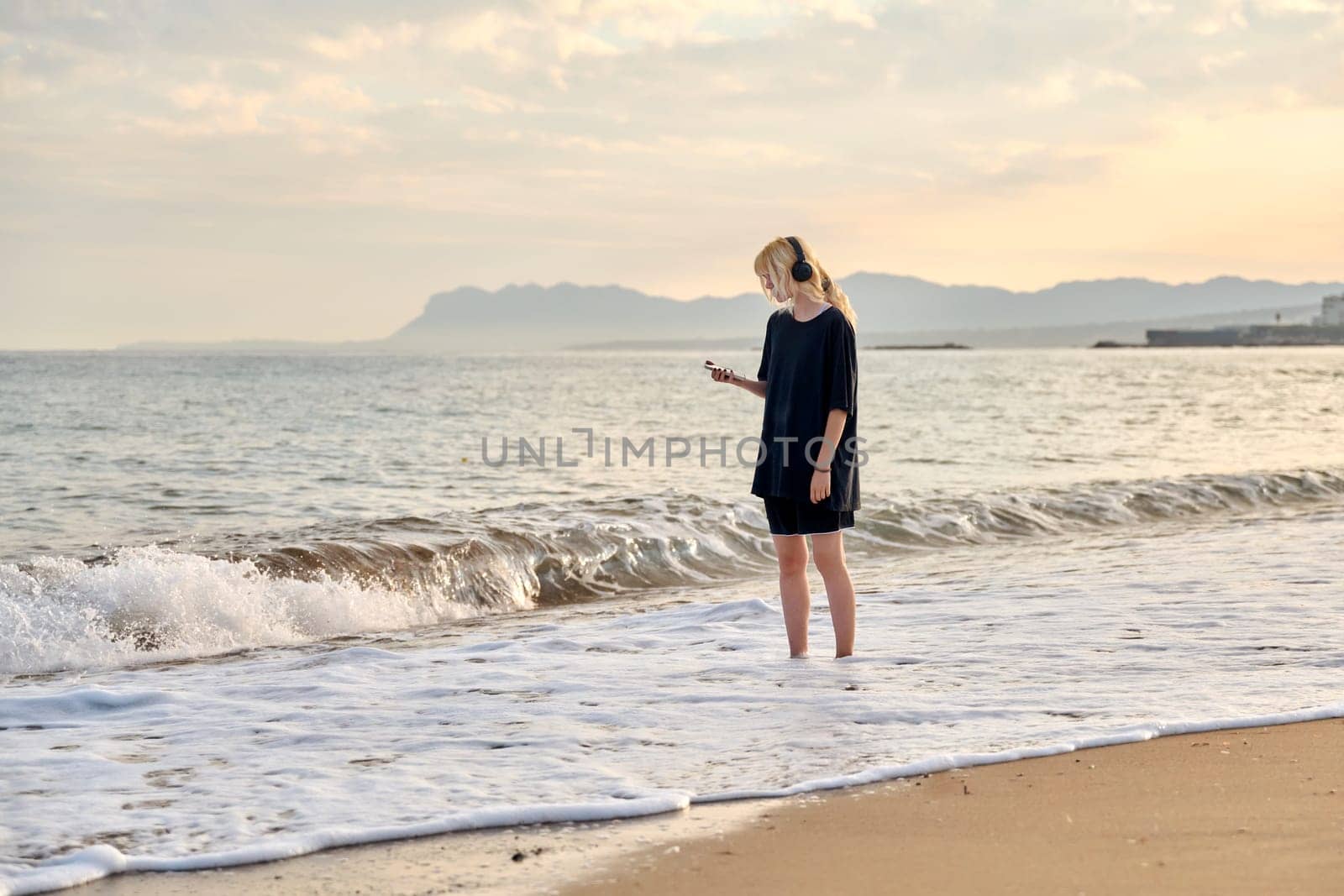 Young teenage female on beach in morning. Hipster teenager in headphones walking along seashore with picturesque colorful sea nature. Summer sunrise, freedom vacation beauty youth concept