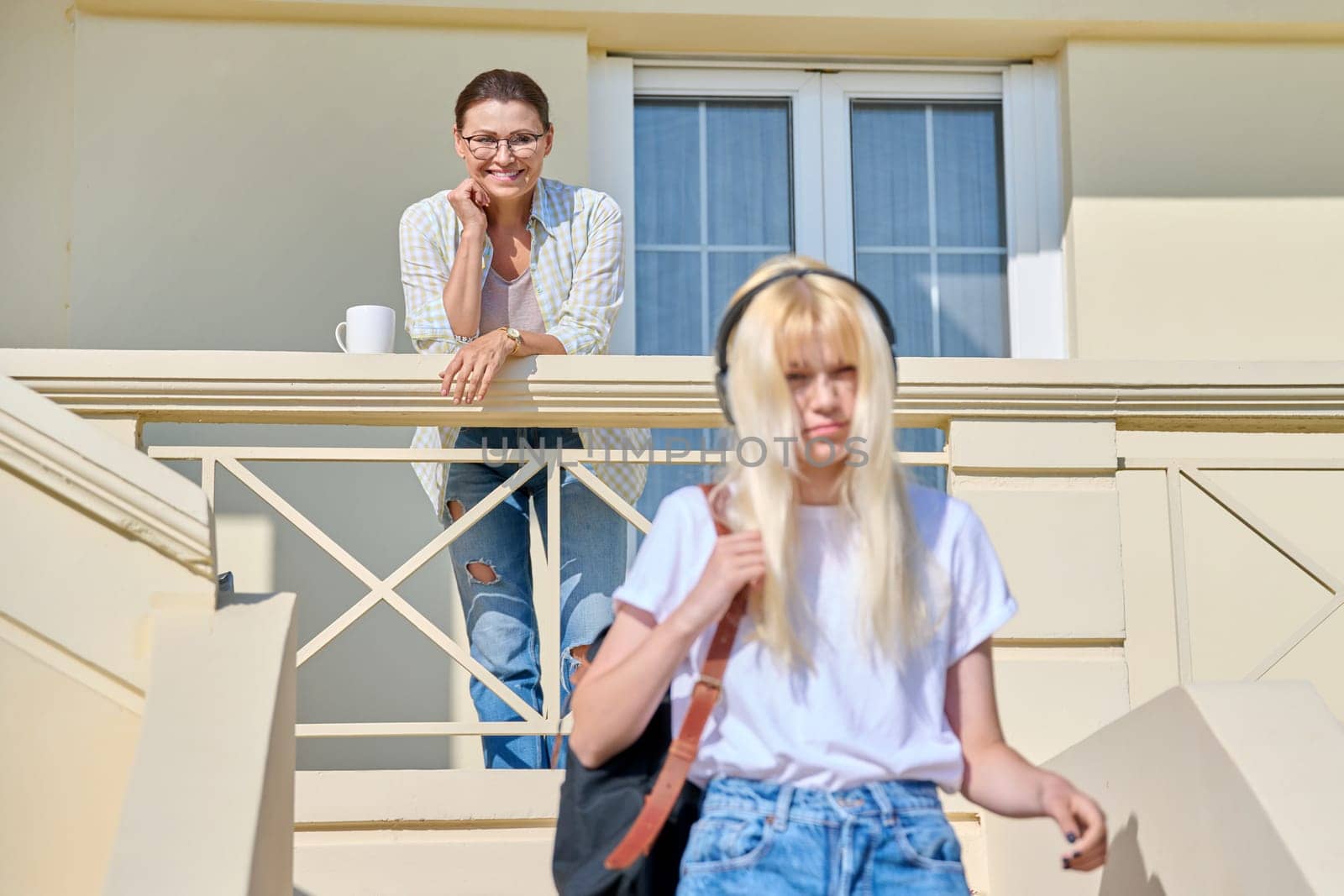Middle-aged mother seeing off her teenage daughter on the porch of the house. Girl student in headphones with a backpack walking up the stairs. Family, lifestyle, parent and teenager concept