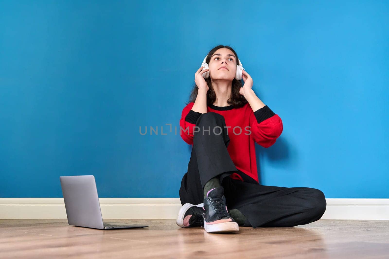 Young teenage guy student in headphones with laptop sitting on the floor on blue background. Lifestyle, youth, technology, leisure, music, education, young people concept