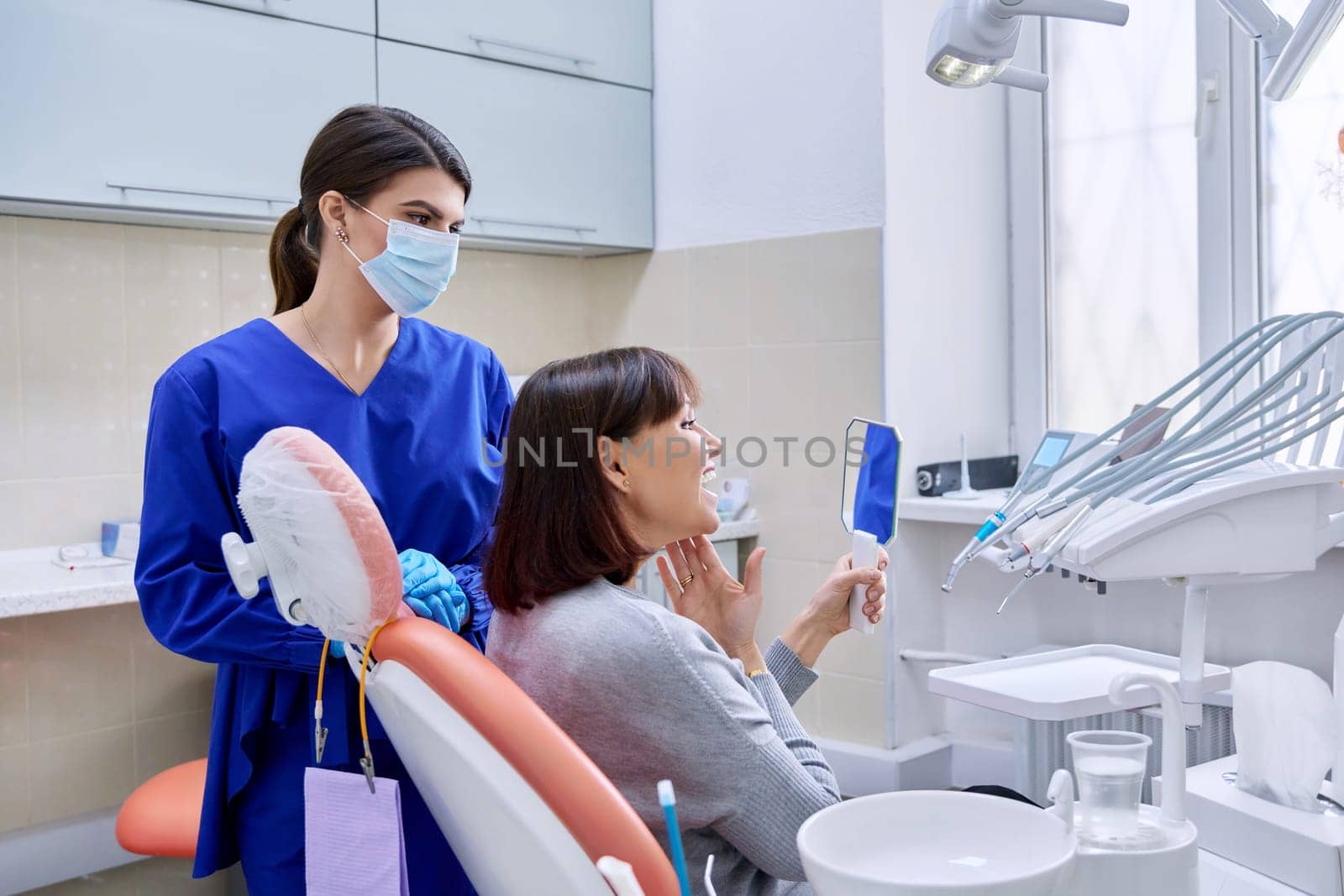 Dentist's office, woman patient looking at her teeth in the mirror. Doctor dentist near the dental chair with tools. Treatment, dental care, prosthetics, orthodontics, dentistry concept