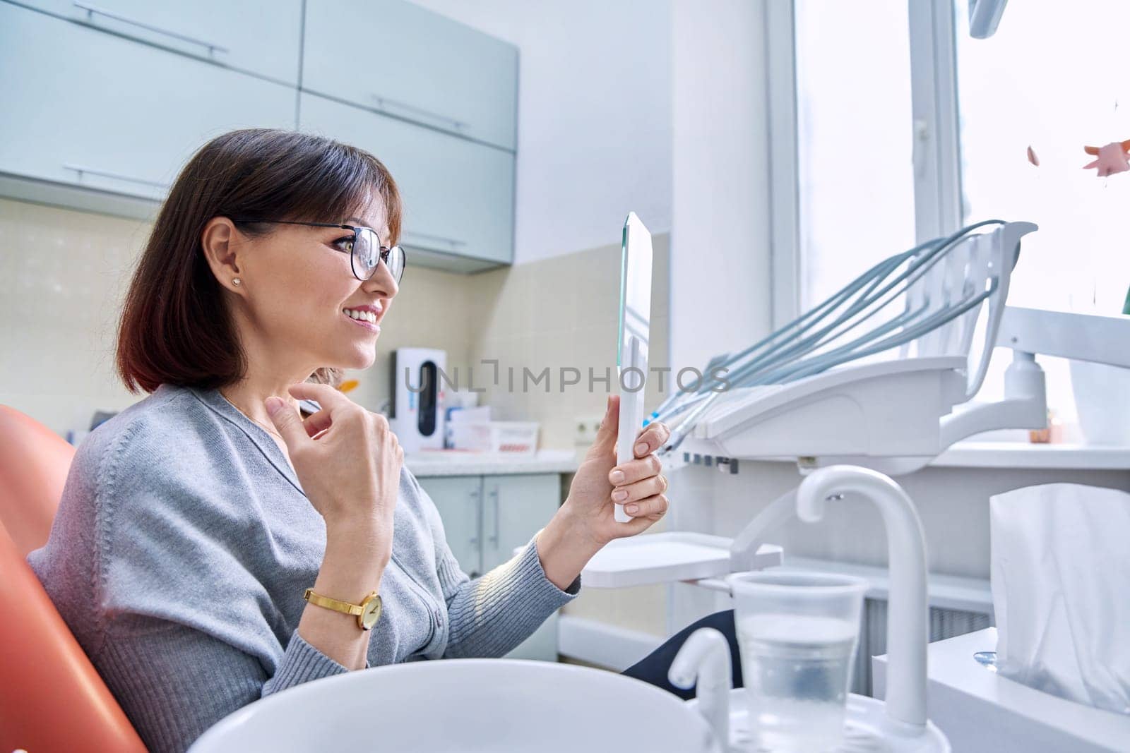 Smiling middle aged woman in dental chair in dentist's office with mirror looking at her teeth. Treatment, therapy, dental care, prosthetics, dentistry concept