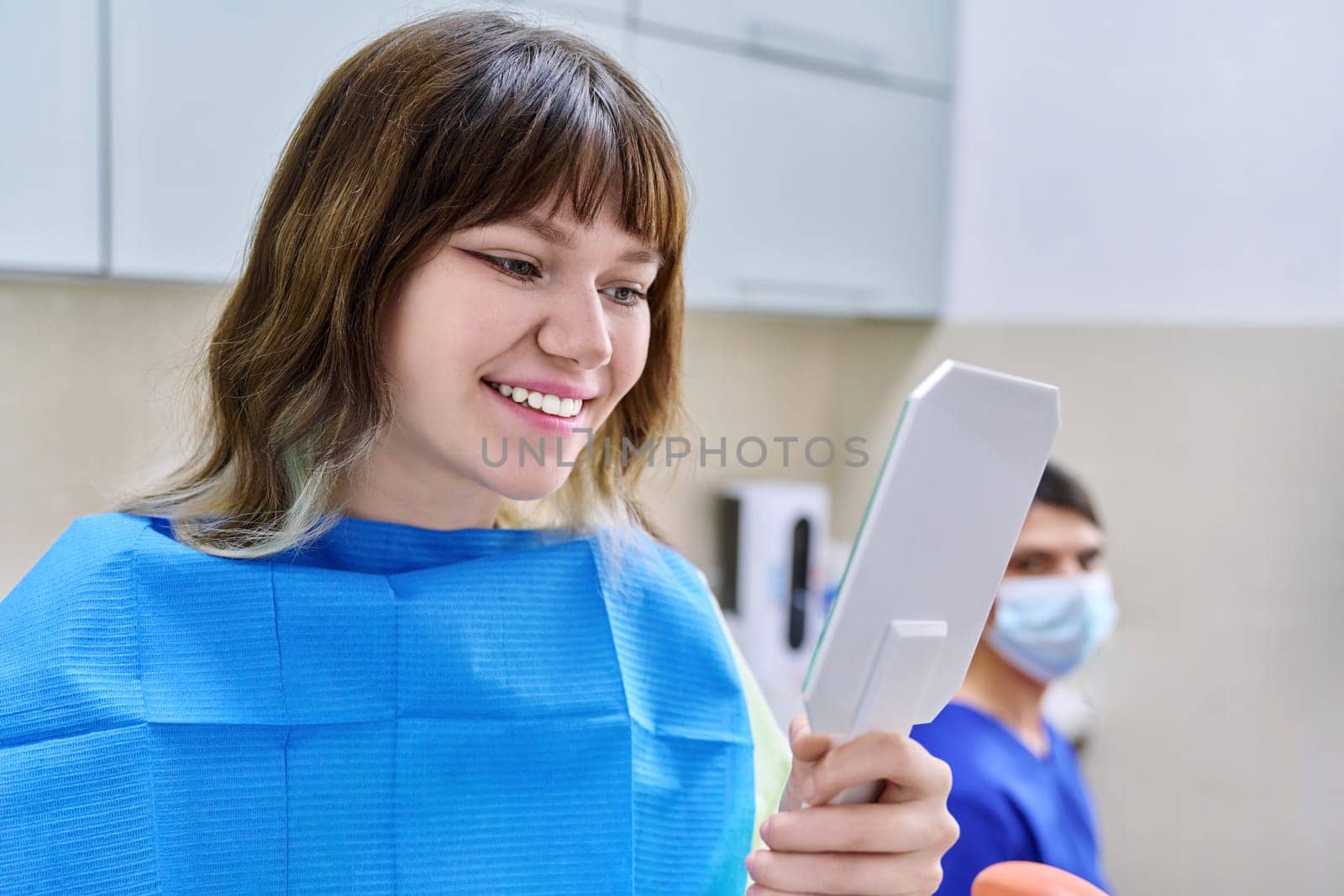 Teenage female looking at healthy teeth in mirror, in dental office, visit to dentist orthodontist. Treatment and care of teeth, teenagers, orthodontics, dentistry concept