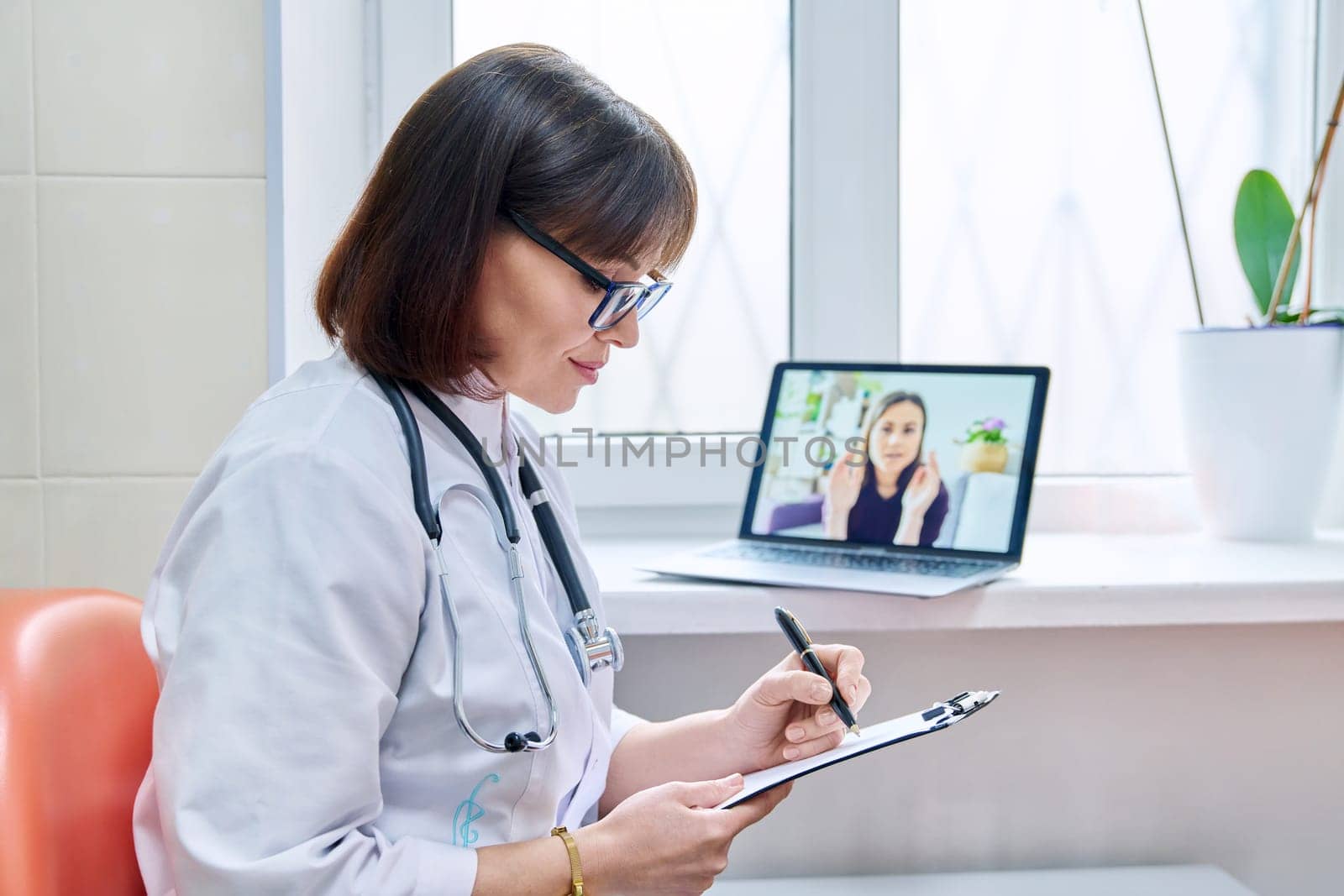 General practitioner doctor talking on videoconference with female patient making notes on file holder. Video call on laptop, online virtual help, remote medical consultation. Medicine technology
