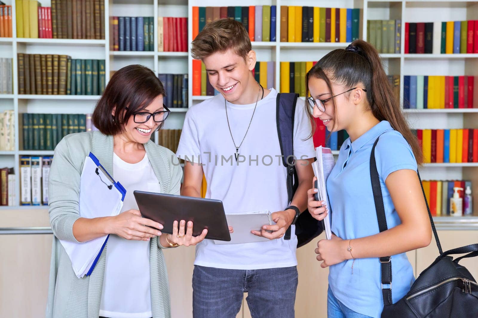 Students teenagers talking with woman teacher mentor, library shelving with books background. High school, education, knowledge, adolescence concept