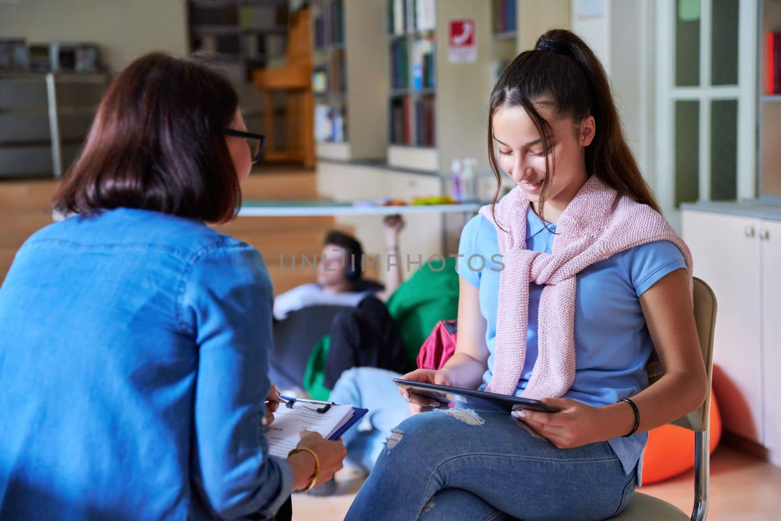 Teenage girl student talking with teacher mentor in the library. Education, knowledge learning, high school, adolescence concept