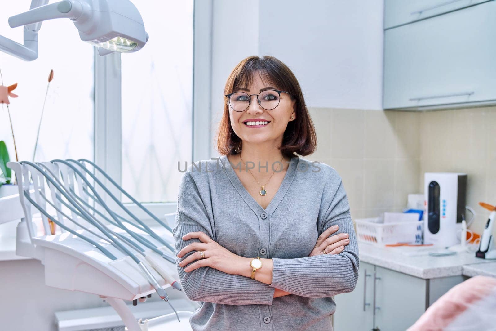 Portrait of smiling mature woman in dental clinic looking at camera. Confident female with crossed arms, dental office interior with equipment. Treatment, therapy, dental care, prosthetics, dentistry