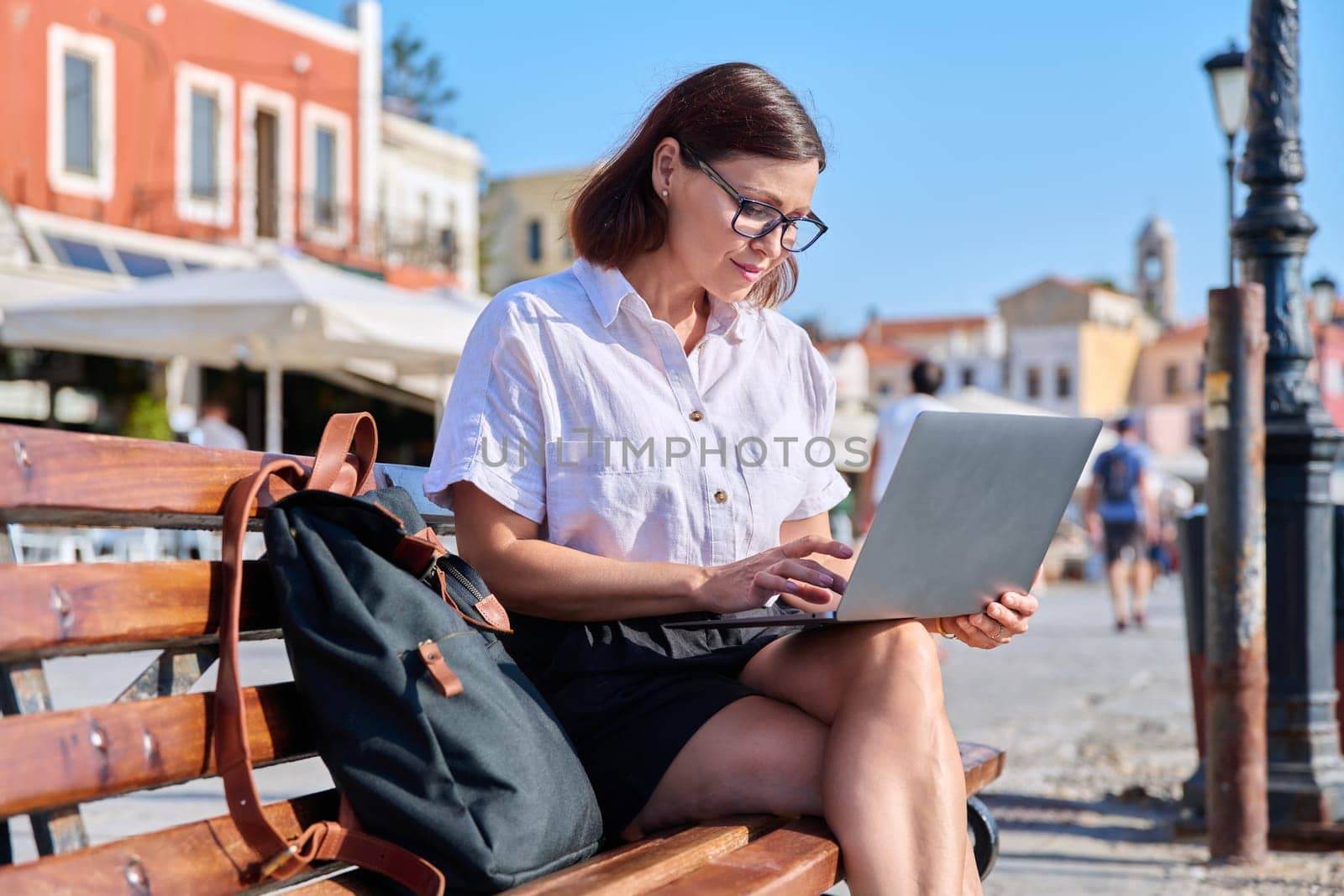 Middle-aged woman with a backpack sitting on a bench in an old tourist city, using a laptop. Freelance, business, technology, mature people concept