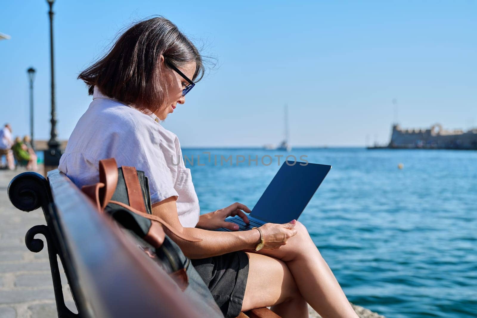 Business mature woman sitting outdoor using laptop. Female 40s with backpack sitting on bench on sea promenade working with laptop. Summer, business travel, freelancing, vacation, people concept