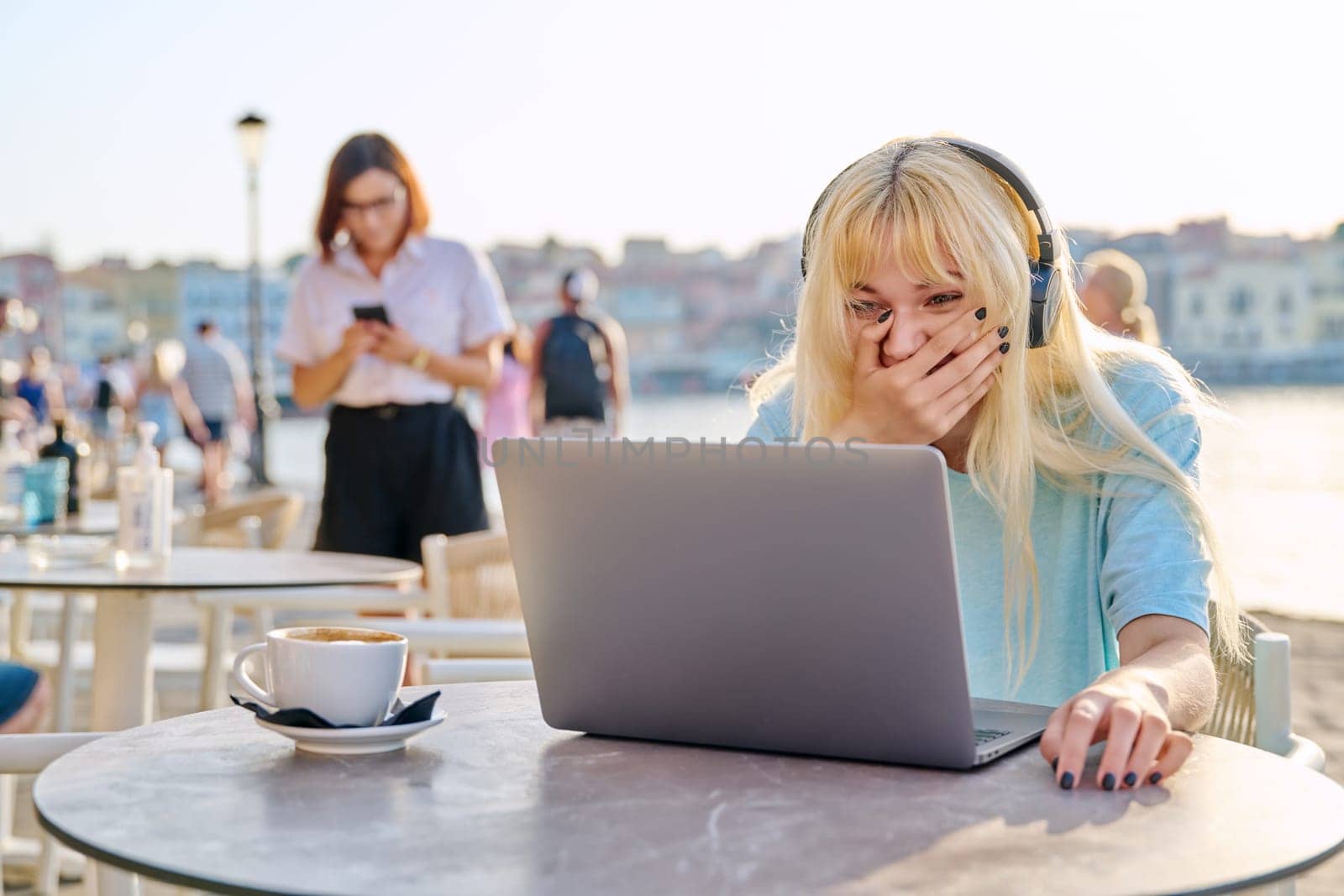 Surprised teenage female with headphones looking at a laptop in an outdoor cafe. Beautiful emotional young blonde on a summer day at a table with a cup of coffee on the seaside promenade.