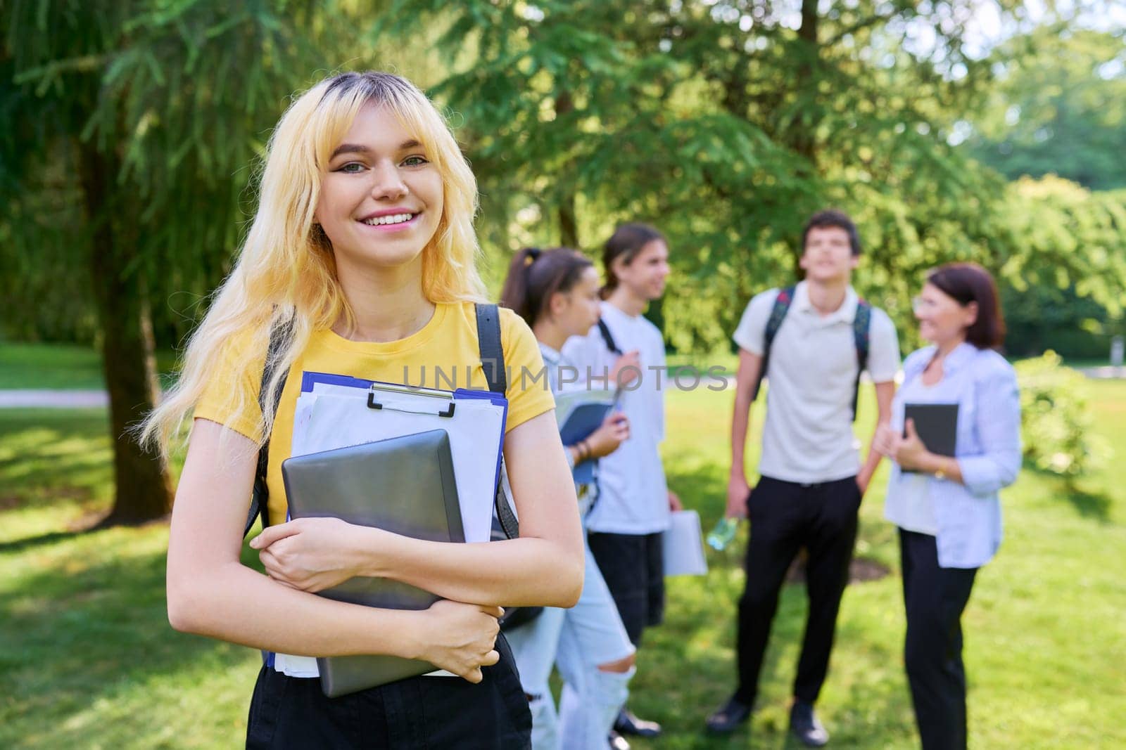 Female student 16, 17 years old with textbooks backpack, group of teenagers talking with teacher in park. Education, teenage students, adolescence, school concept