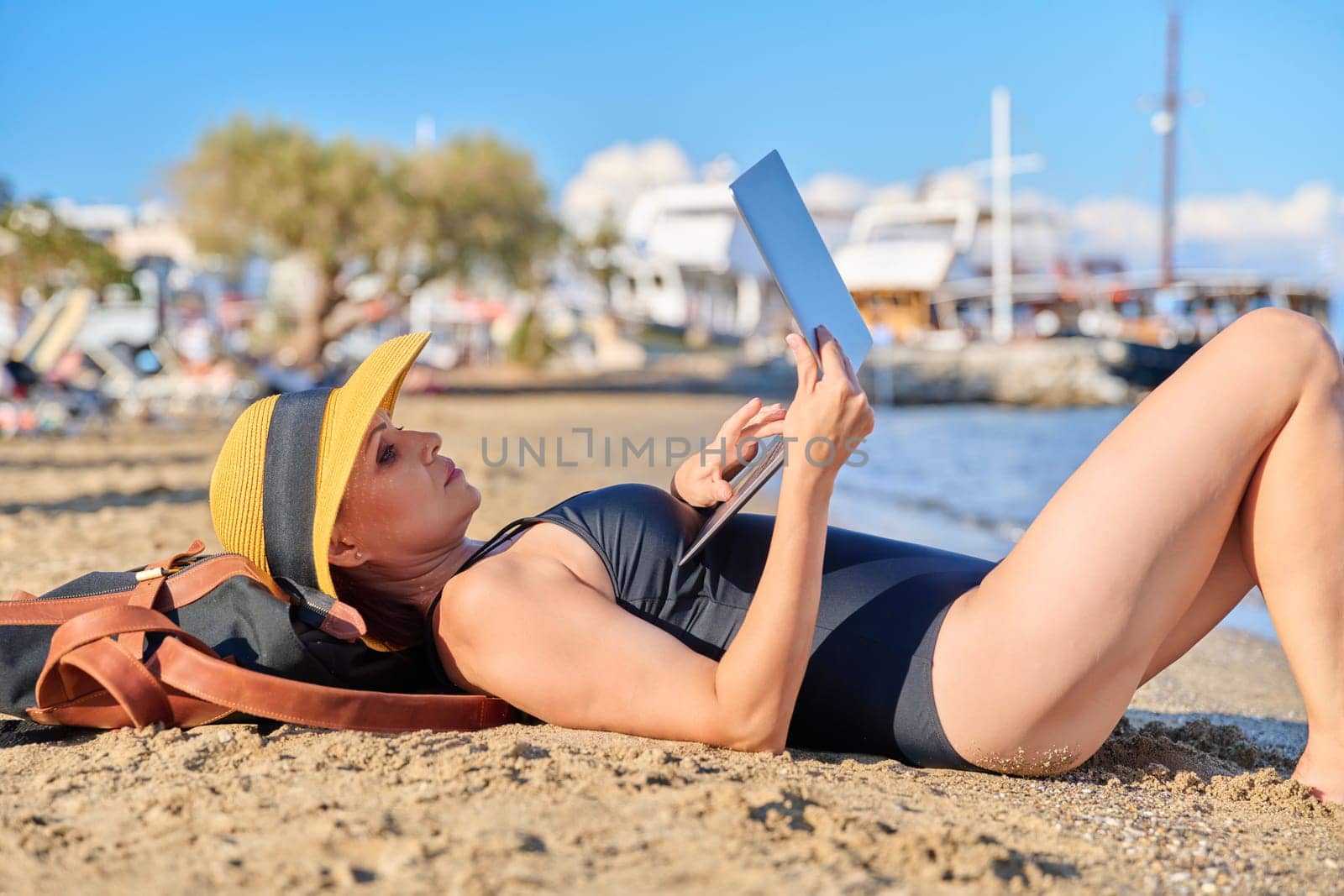 Mature woman sunbathing on beach in hat with laptop, female in swimsuit lying on sand, using laptop. Freelance, remote work, leisure, leisure, relaxation, technology and middle aged people concept