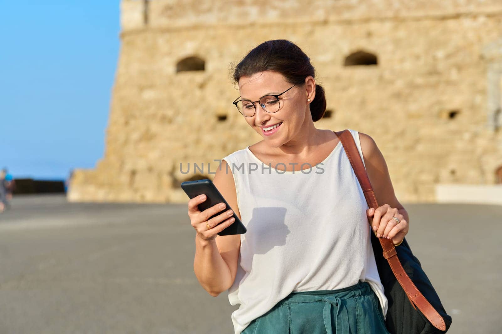 Beautiful mature woman walking with smartphone in hand, female with a backpack in the old historical city. Active lifestyle, travel, tourism, middle aged people concept