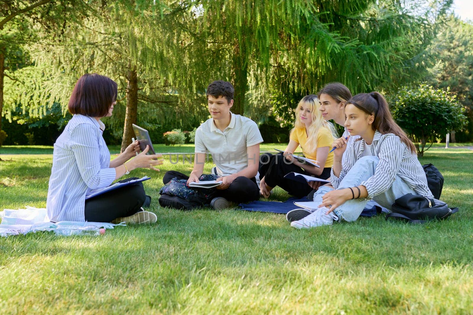 Outdoor, group of students with female teacher. Teenagers and mentor teacher talking sitting on grass in college park. Back to school, back to college, high school, education, teenagers concept