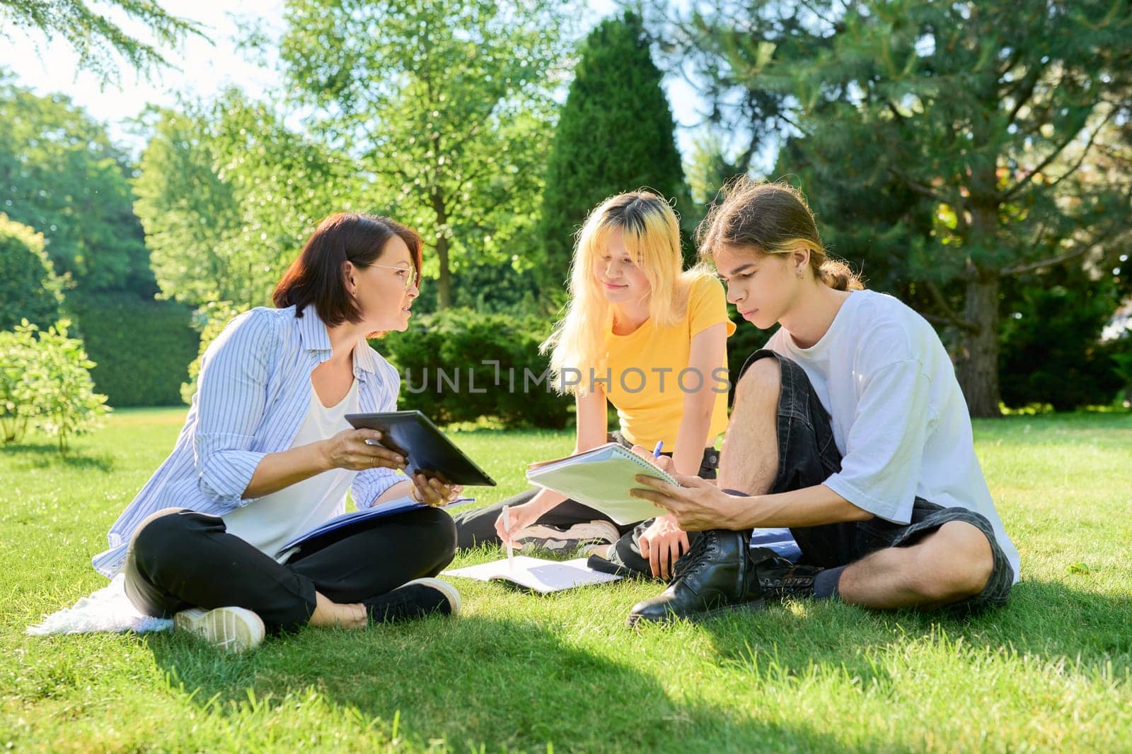 School teacher, psychologist, social worker talking to teenagers, sitting on grass by VH-studio